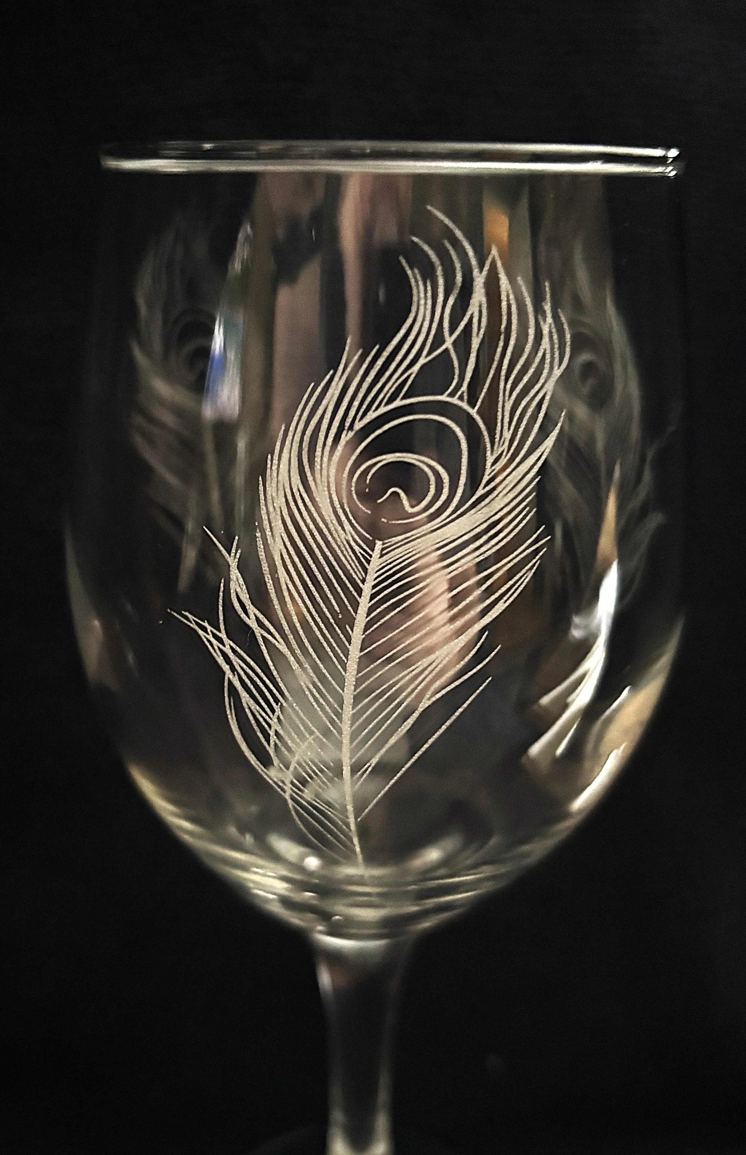 A clear wine glass with an engraved peacock feather design etched on its surface, standing against a dark background.