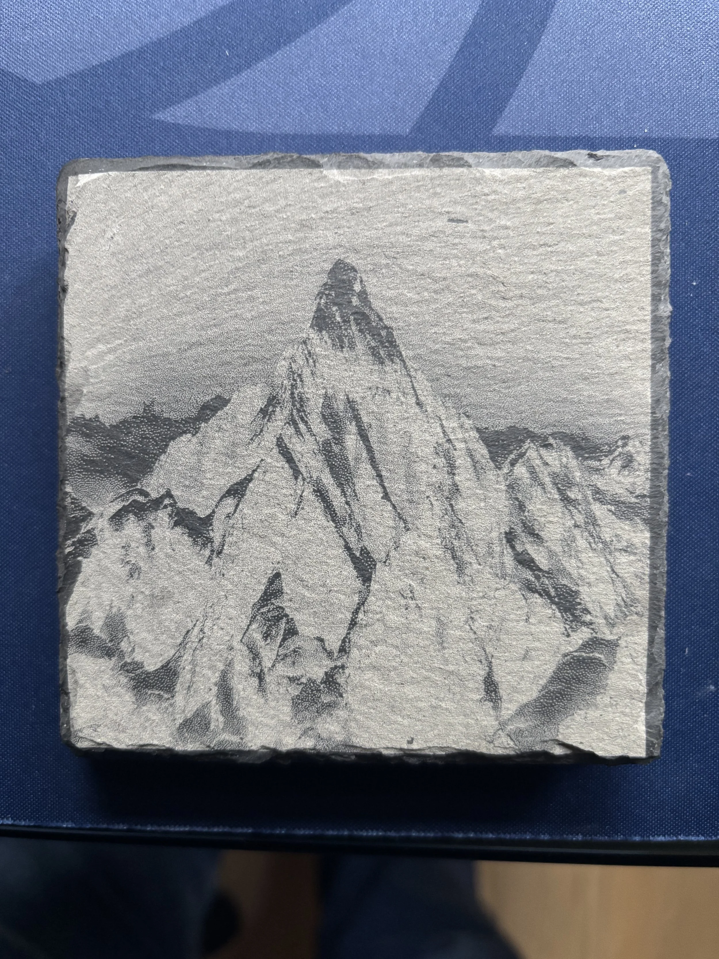 A silver-colored, textured metal block with an engraved image of a mountain peak and surrounding mountains.