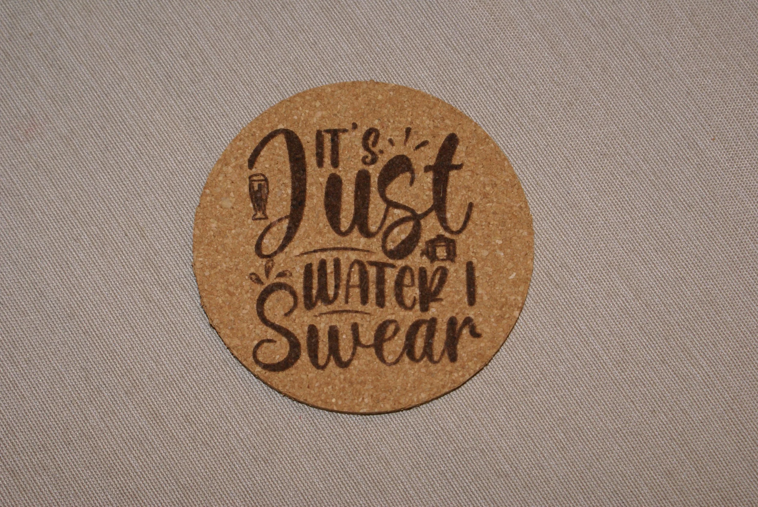 A round coaster with the phrase "It's just. Watch. Swer" written in a decorative font on a light fabric background.