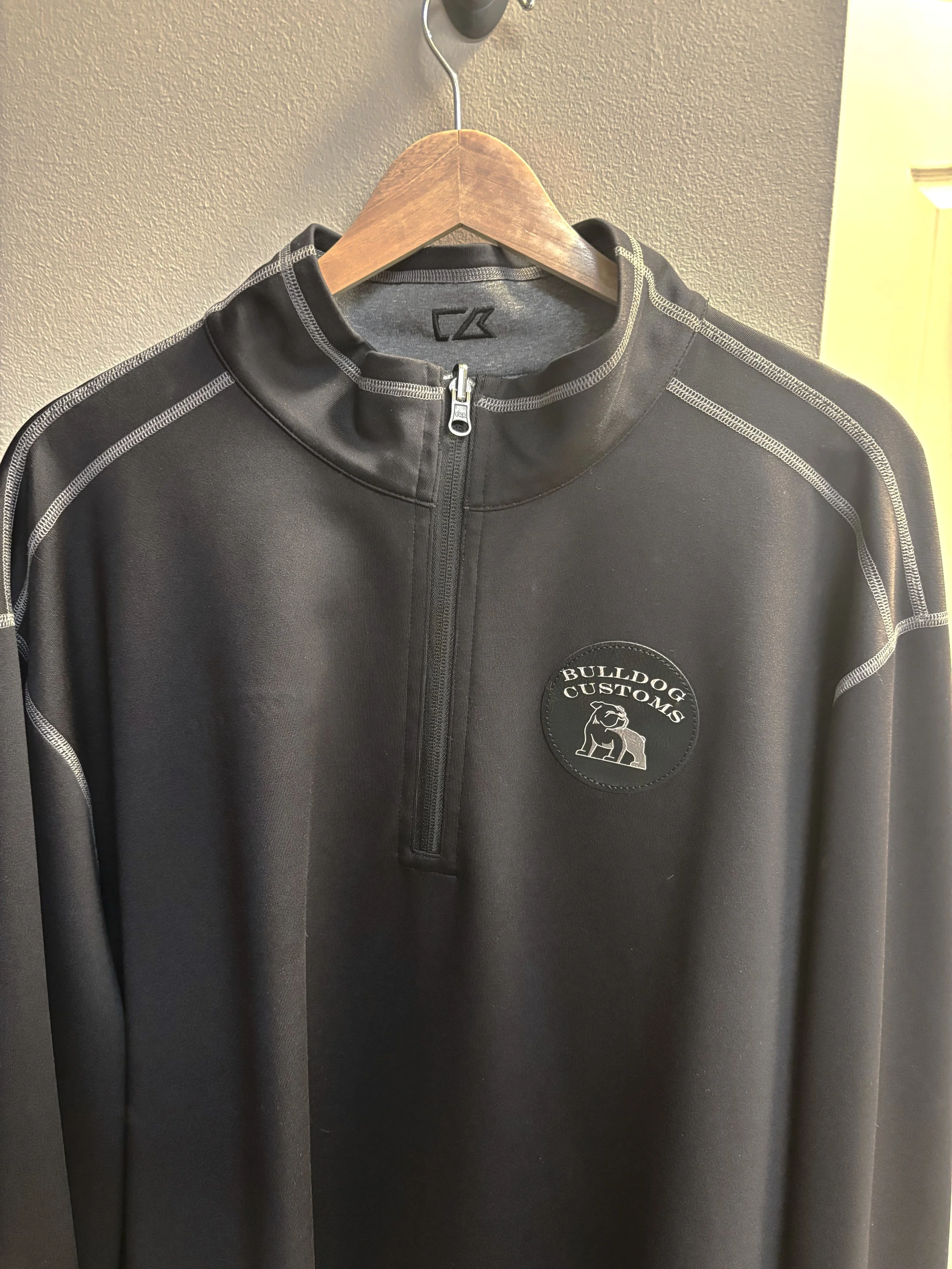 Black athletic jacket with gray stitching and a logo patch on the chest that reads 'Bulldog Customs' with a bulldog graphic will a ban. The jacket hangs on a wooden hanger on a wall.