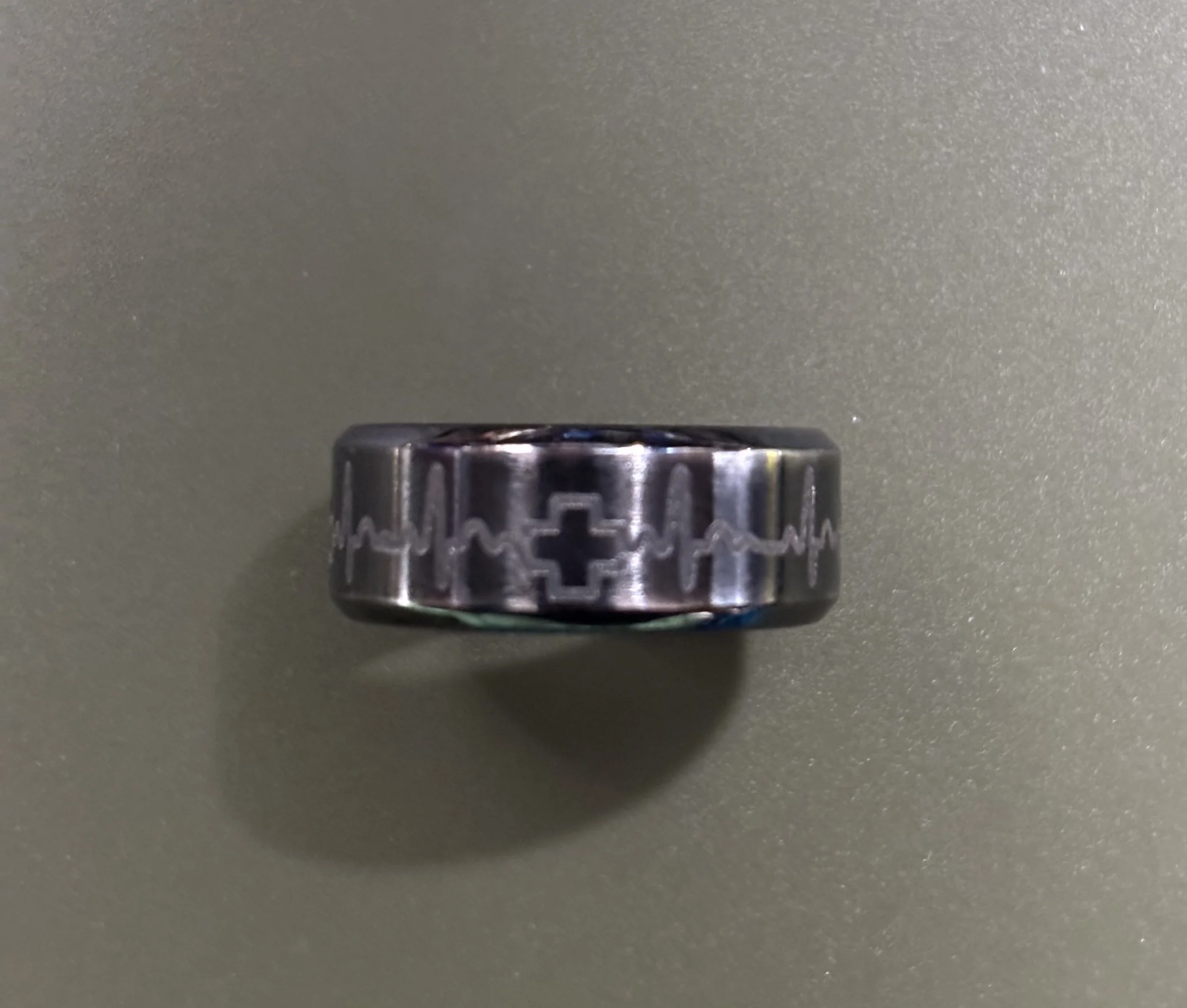 A metallic ring with a cross symbol and a heartbeat line engraved around it, placed on a gray surface.