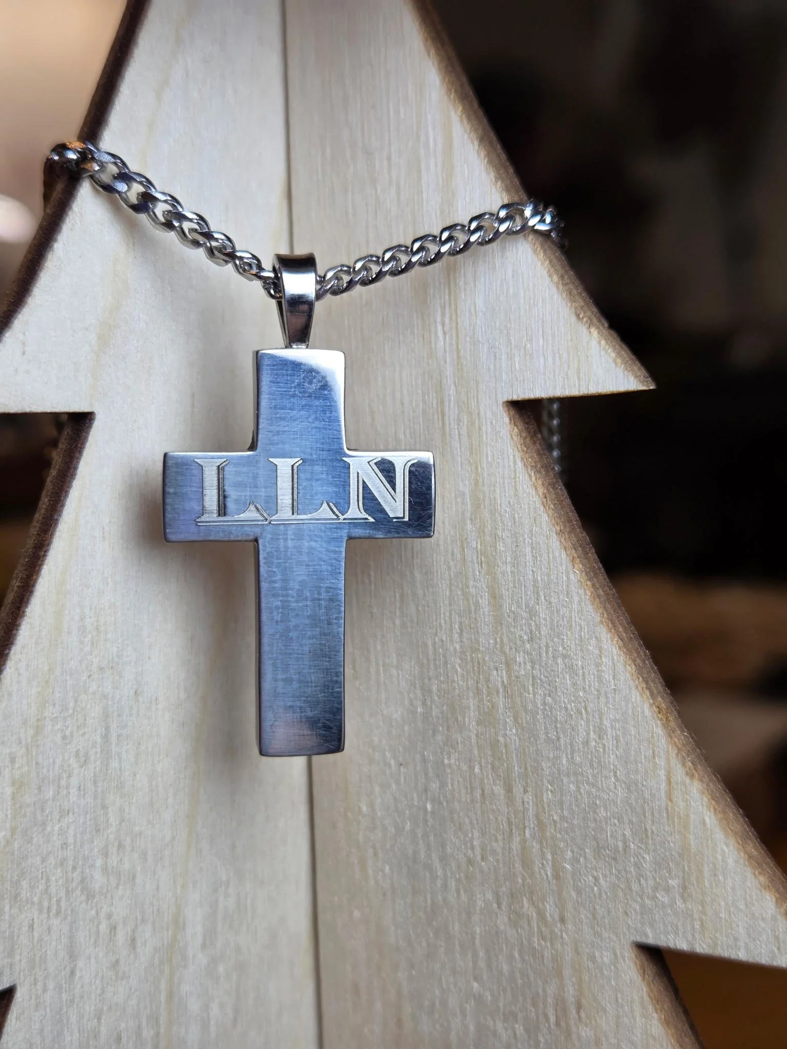 A silver cross necklace with the initials 'LLN' engraved on it, hanging from a silver chain, displayed against a light-colored wooden star-shaped background.