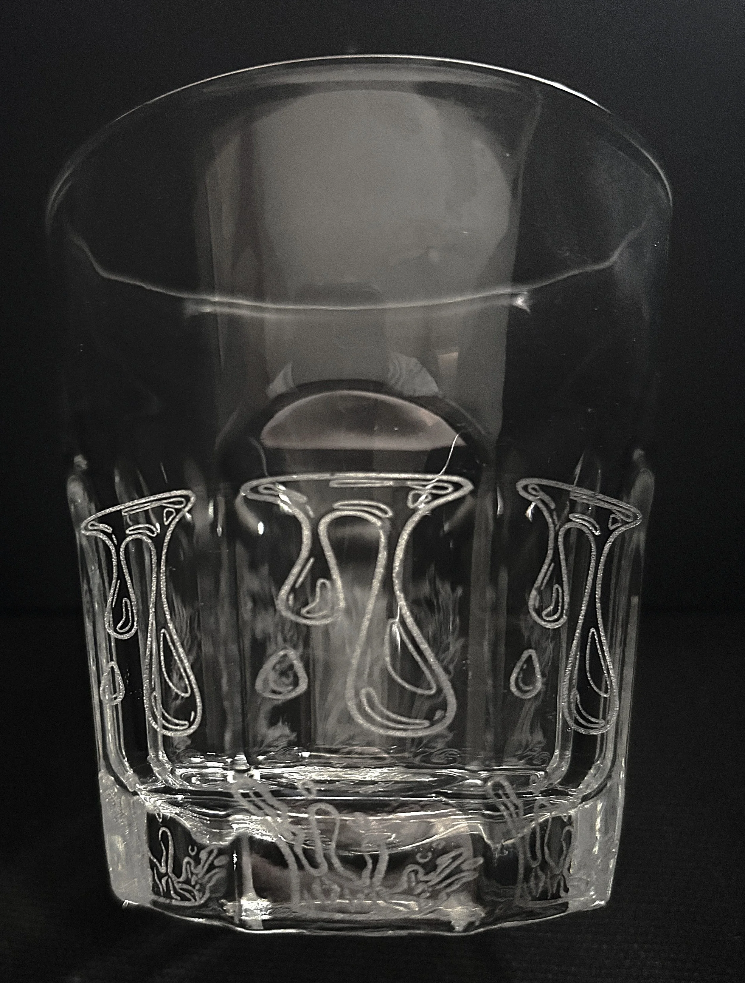 A clear glass tumbler with intricate etched patterns and drops, set against a black background.