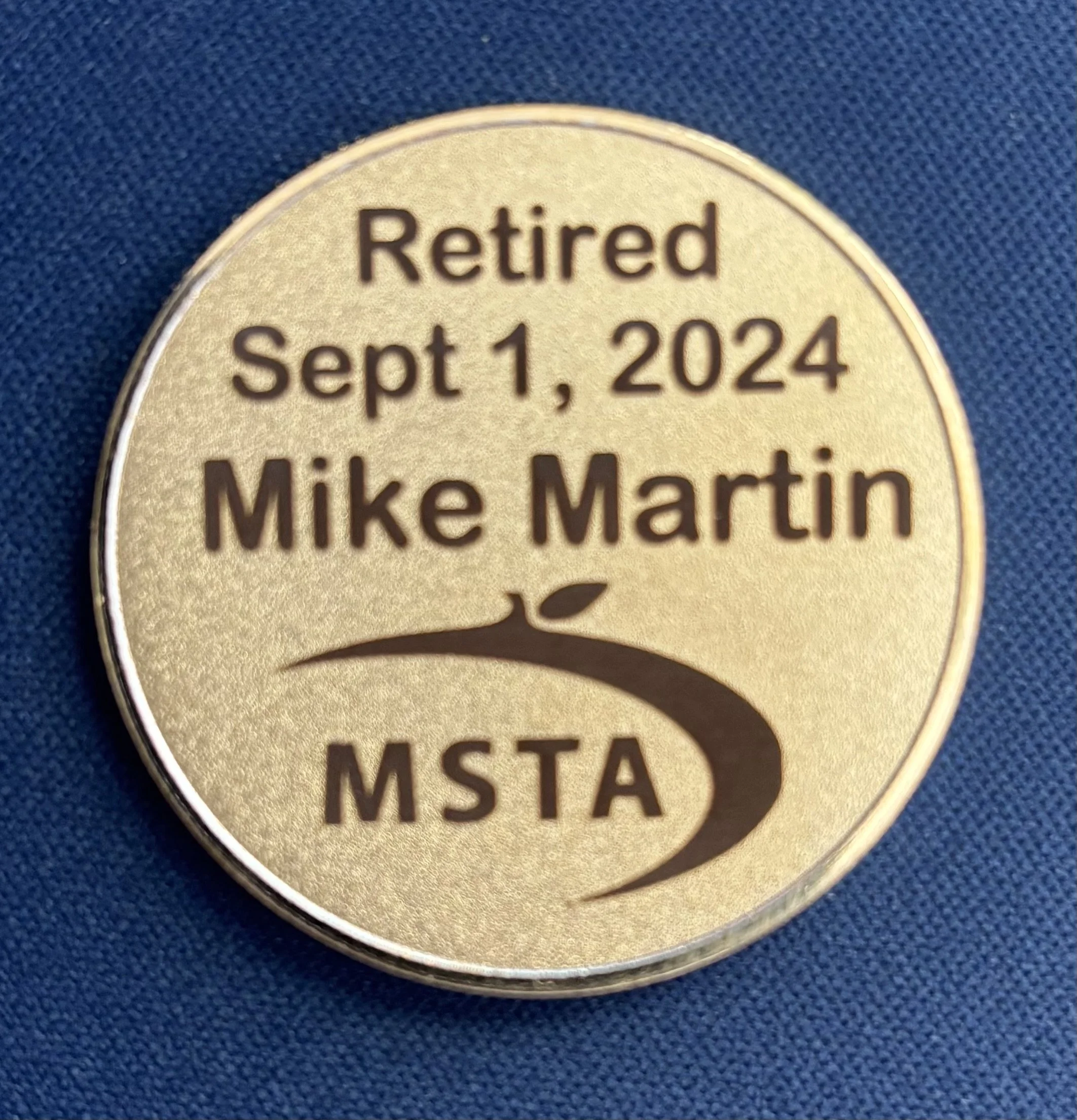A circular badge with a gold background and black text that reads "Retired Sept 1, 2024 Mike Martin" and features a small, curved symbol resembling a plant sprout above the letters "MSTA" on a dark blue fabric background.