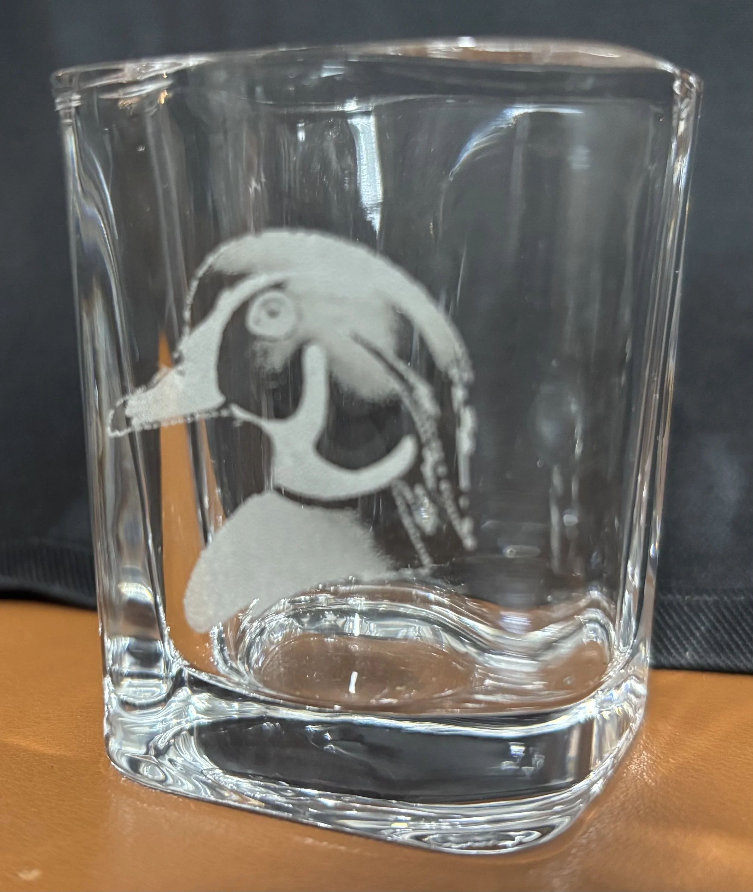 A clear glass with a frosted image of a dog's face etched on it, placed on an orange surface with a black background.