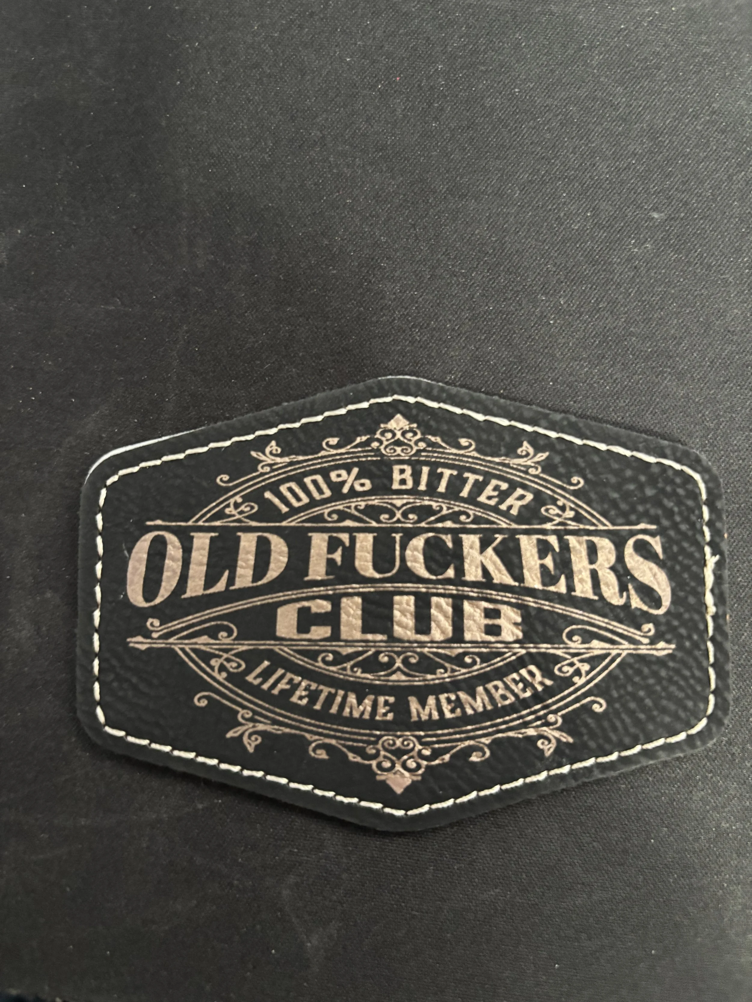 A black patch with white decorative border and text that reads '100% Bitter Old F**kers Club Lifetime Member'.