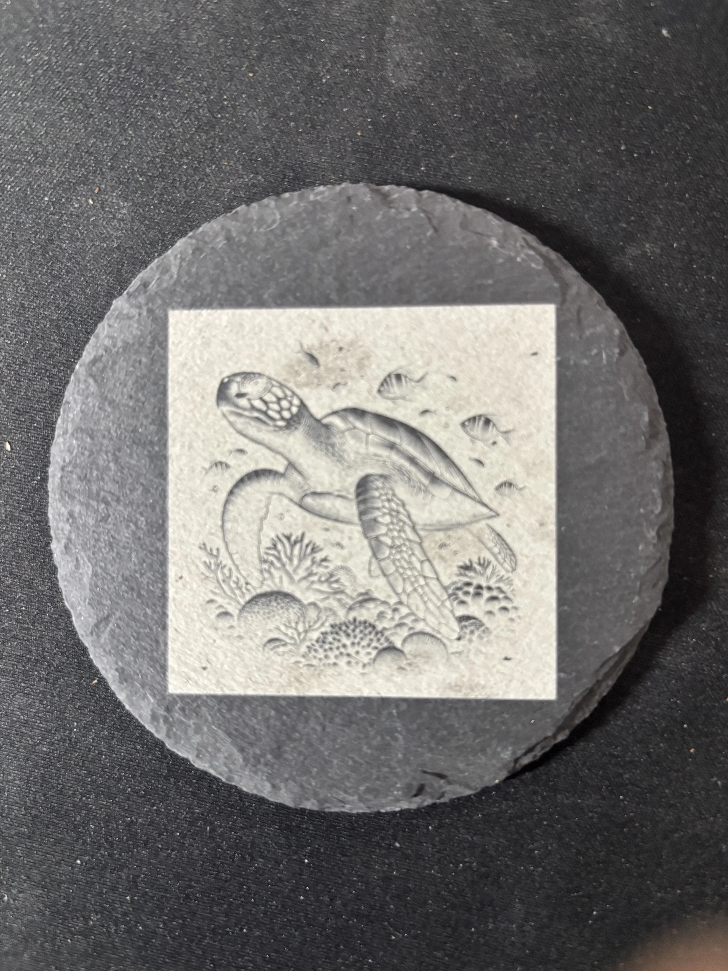 A round, black stone with a gray and white drawing of a turtle swimming underwater, surrounded by small fish and underwater plants.