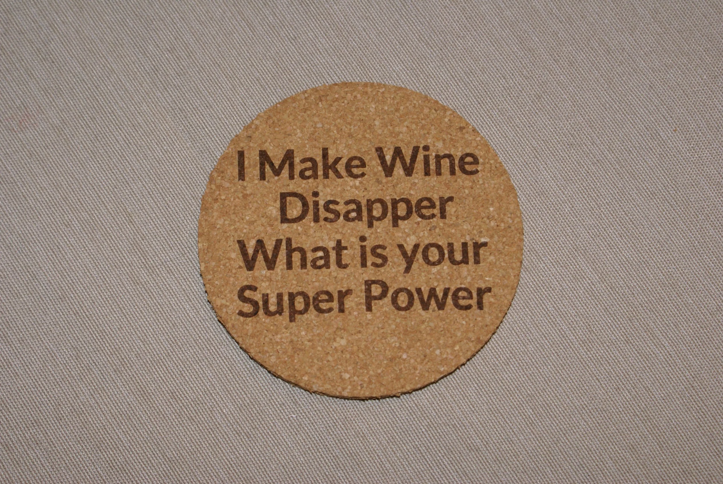 Round cork coaster with the words 'I Make Wine Disappear What is your Super Power' printed on it, placed on a light-colored fabric surface.