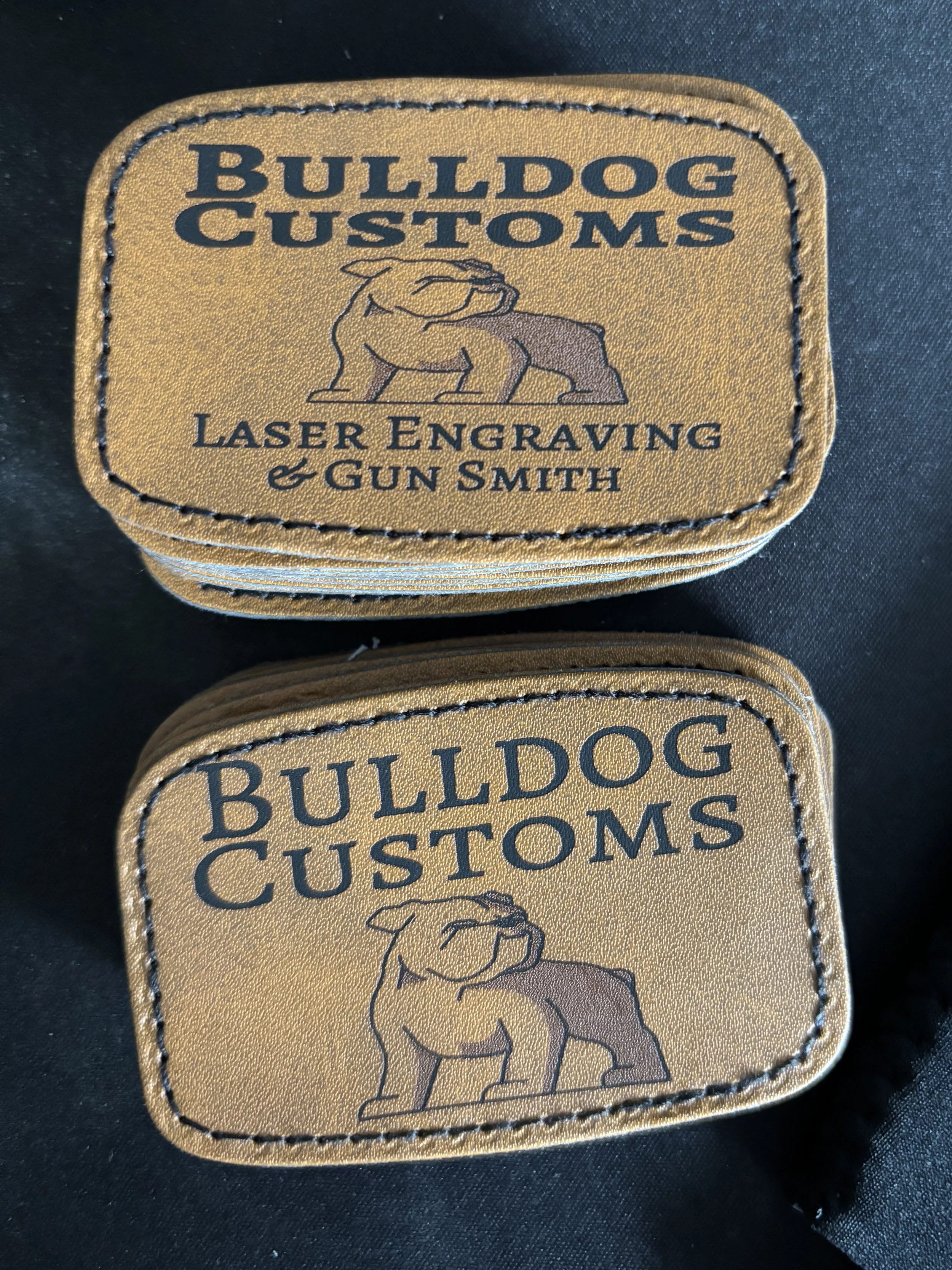 Two tan patches with black stitching, featuring a bulldog logo and the text 'Bulldog Customs Laser Engraving & Gun Smith' on a black fabric background.