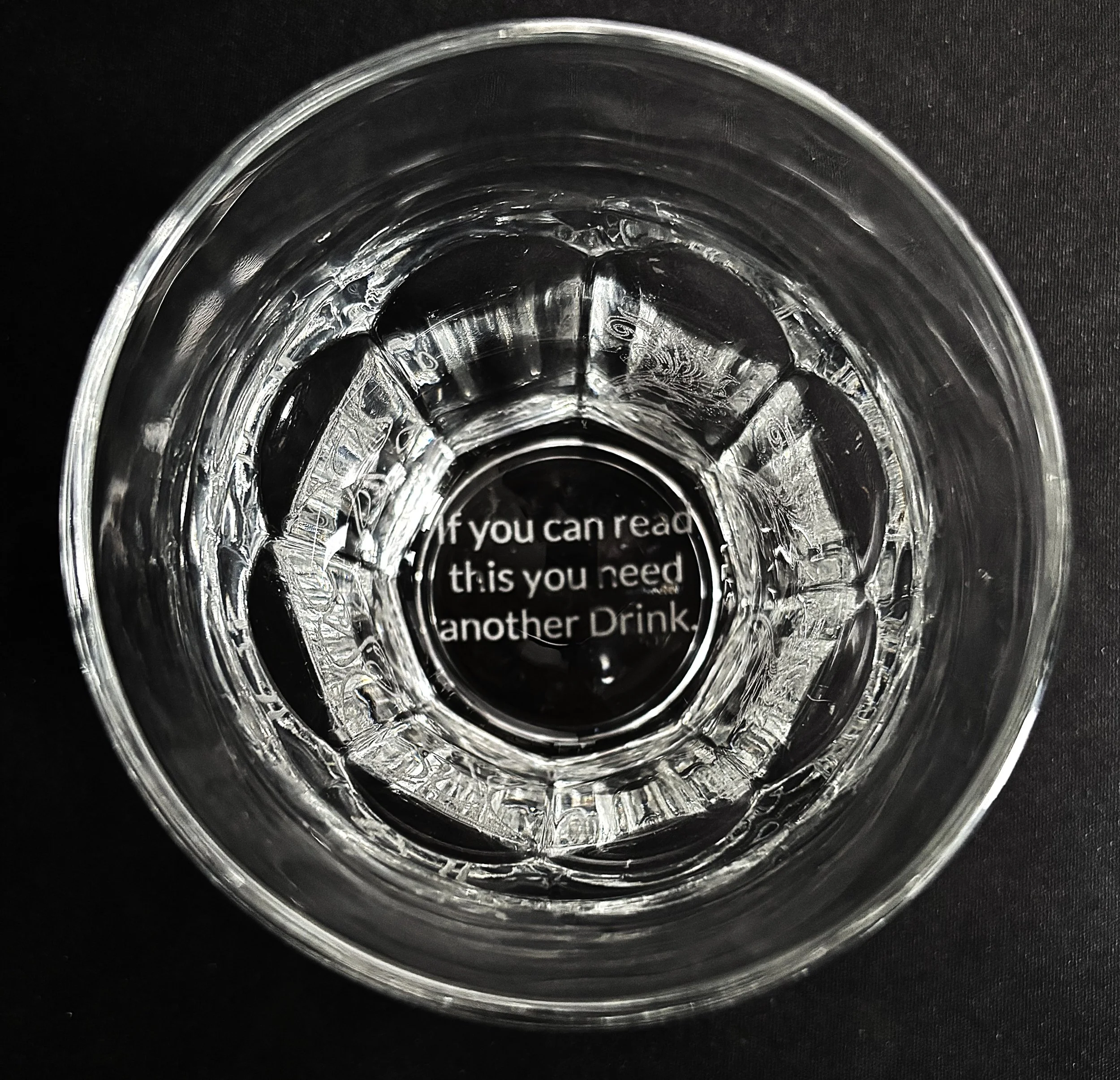 Top-down view of a clear glass with water, focusing on the reflection and the message inside the bottom saying, 'If you can read this you need another Drink'.