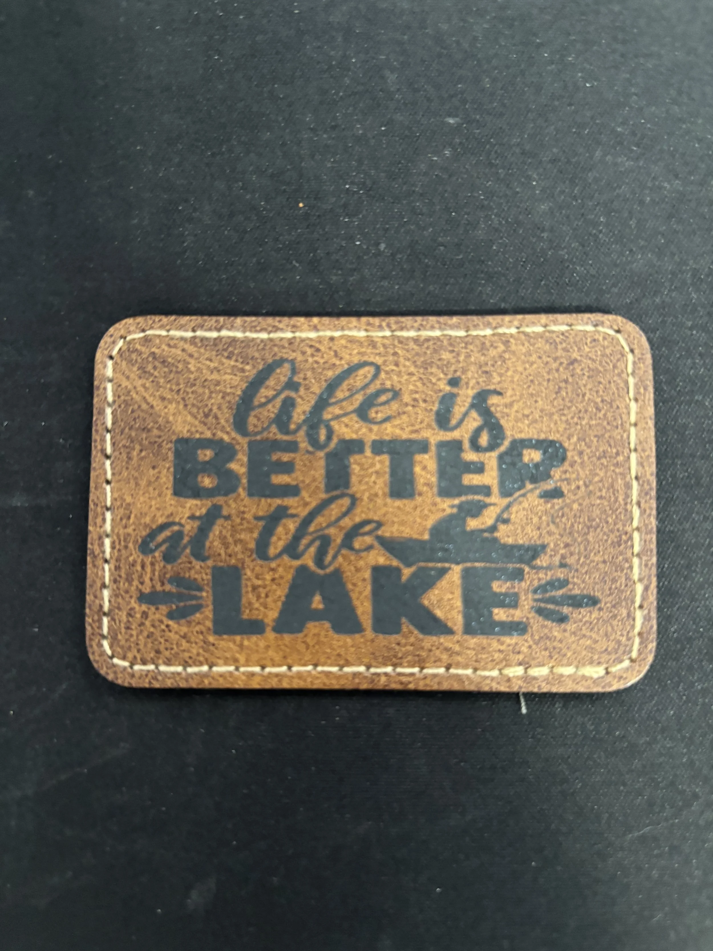 Leather patch with the quote 'life is better at the lakes' on a dark fabric background.