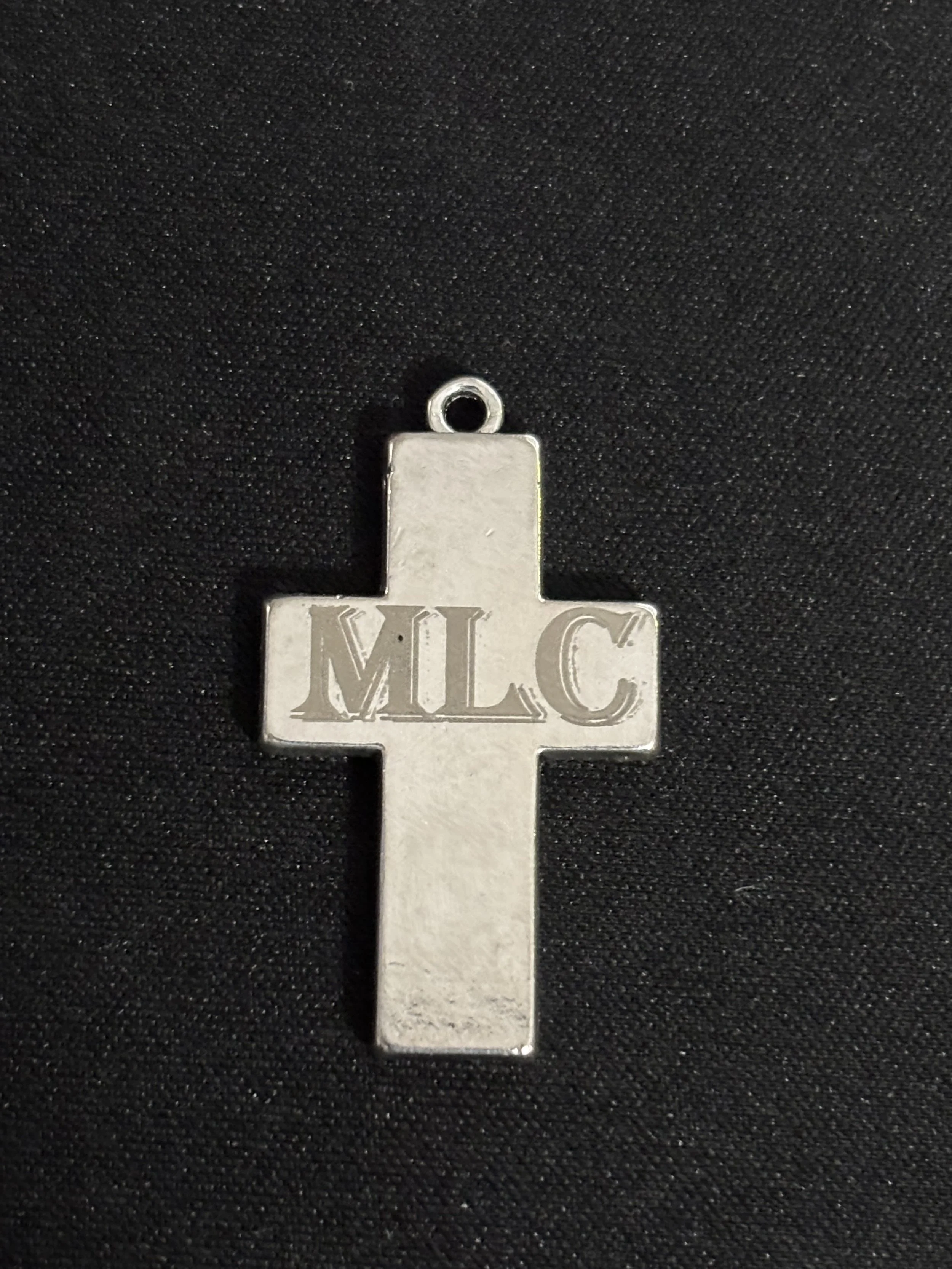 A silver-colored cross-shaped pendant with the initials 'MLC' engraved in the center, lying on a black fabric background.