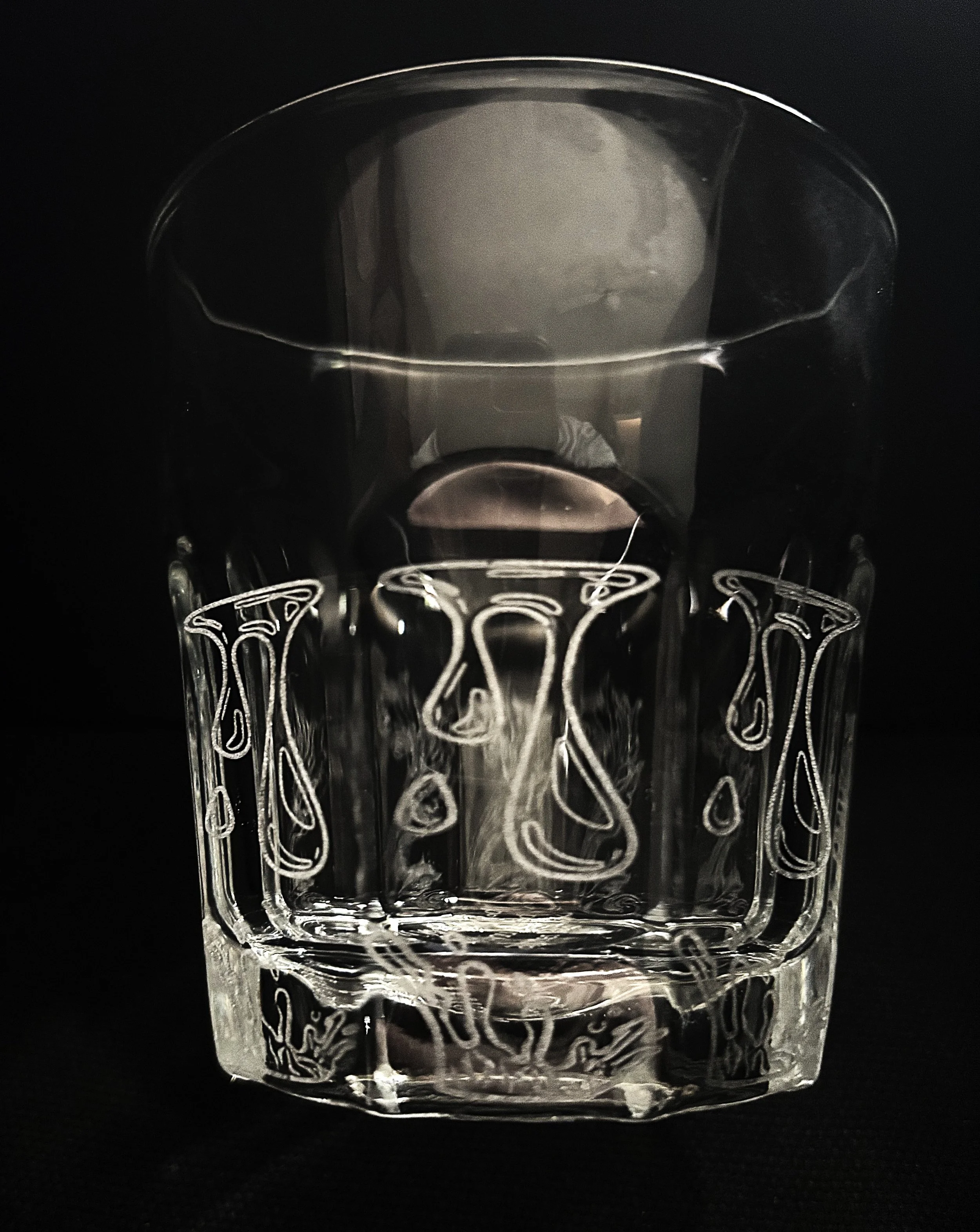 A clear glass tumbler with intricate etched patterns, placed against a black background.