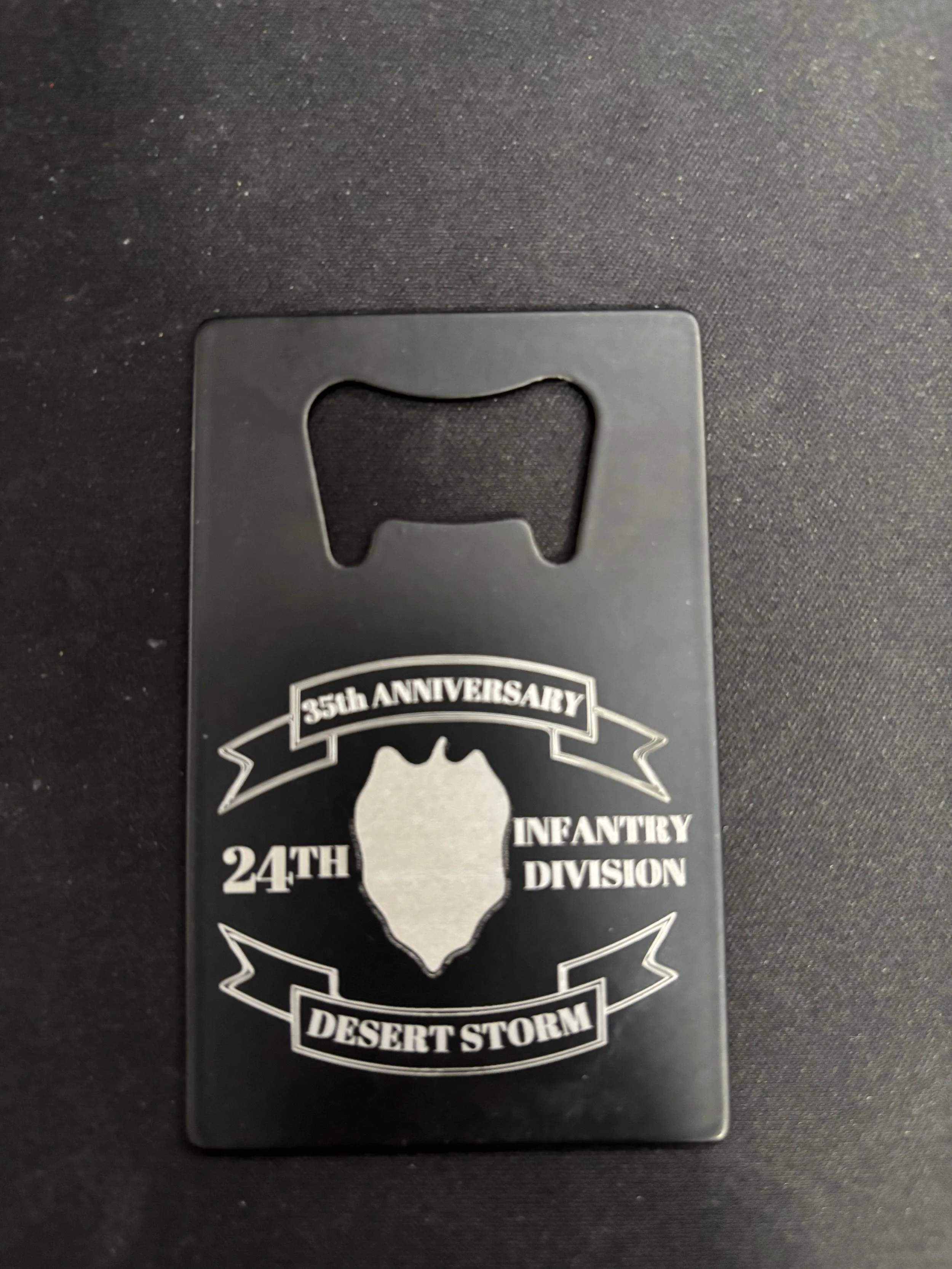 Black bottle opener features an emblem with a shield shape in the center, surrounded by banners that read '35th Anniversary,' '24th Infantry Division,' and 'Desert Storm.'