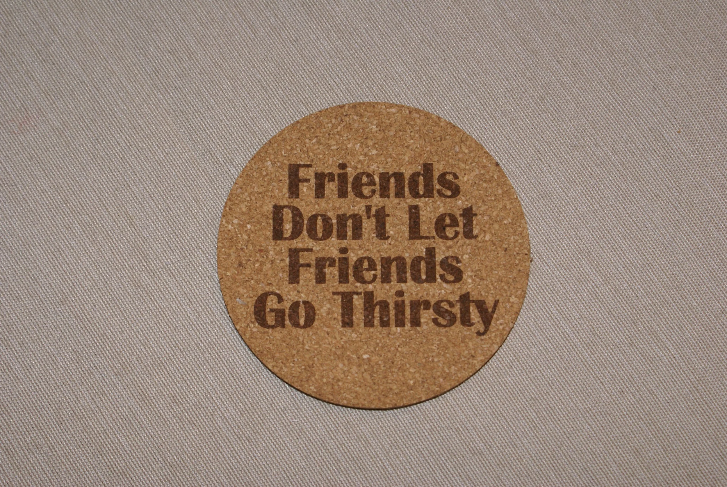 Round cork coaster with the humorous message: "Friends Don't Let Friends Go Thirsty" in brown text on a light-colored fabric background.