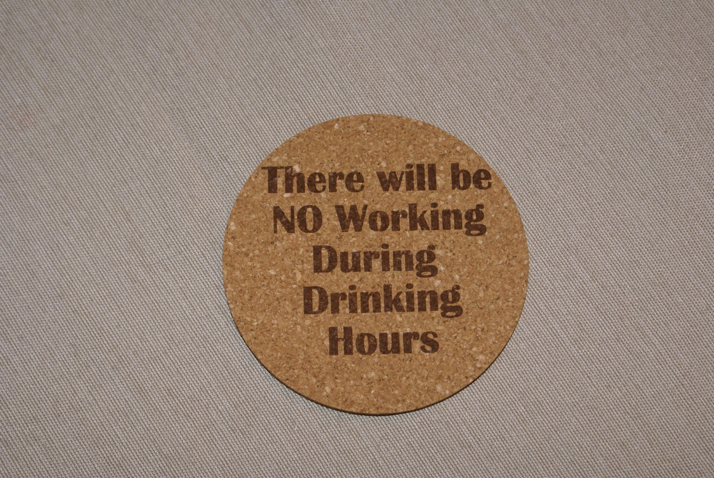 A round cork coaster with text stating 'There will be NO Working During Drinking Hours' on a light gray surface.