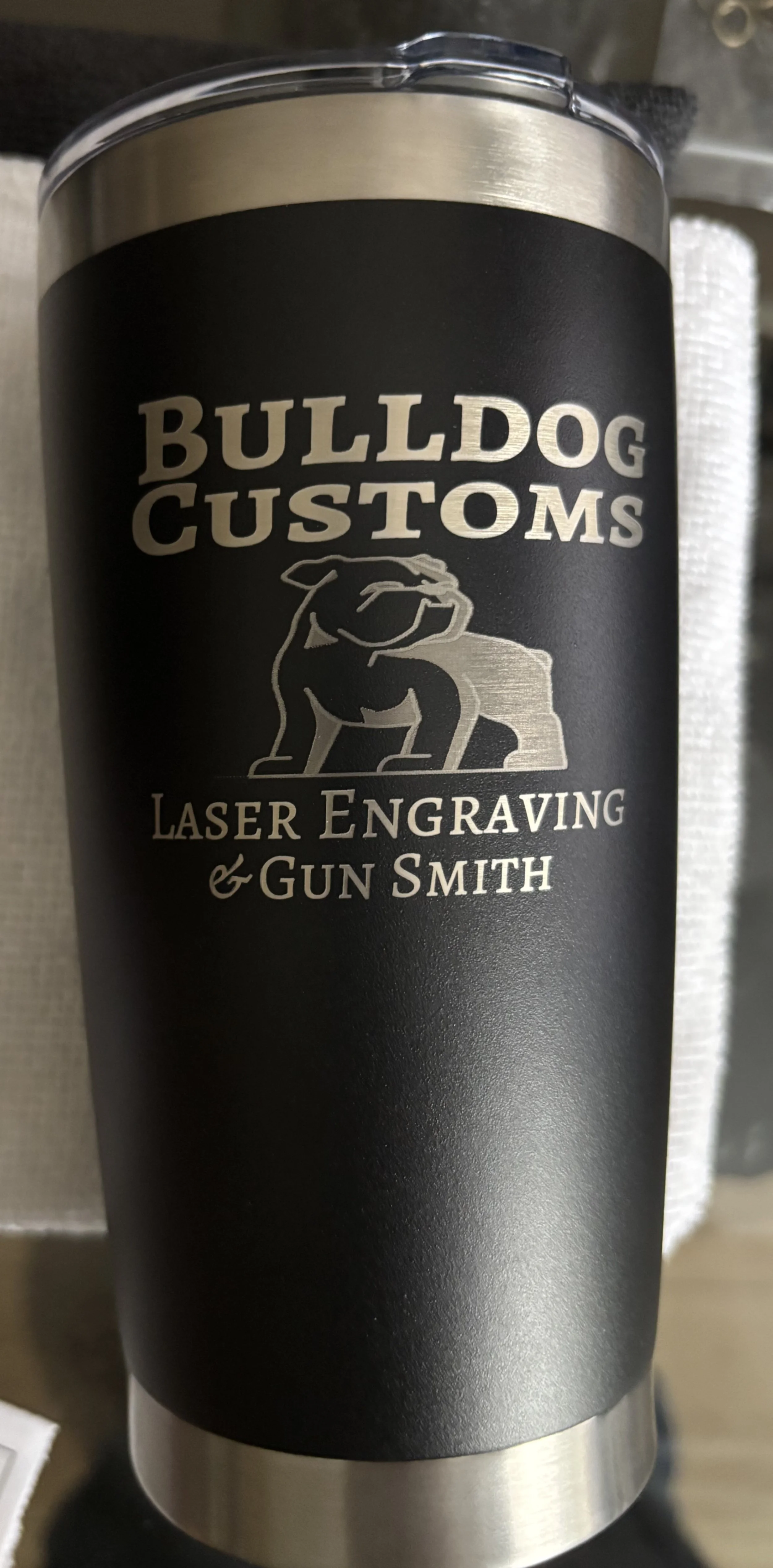 Black travel mug with silver top and bottom, featuring white text and a graphic of a bulldog on a rock. The text reads 'Bulldog Customs Laser Engraving & Gun Smith.'