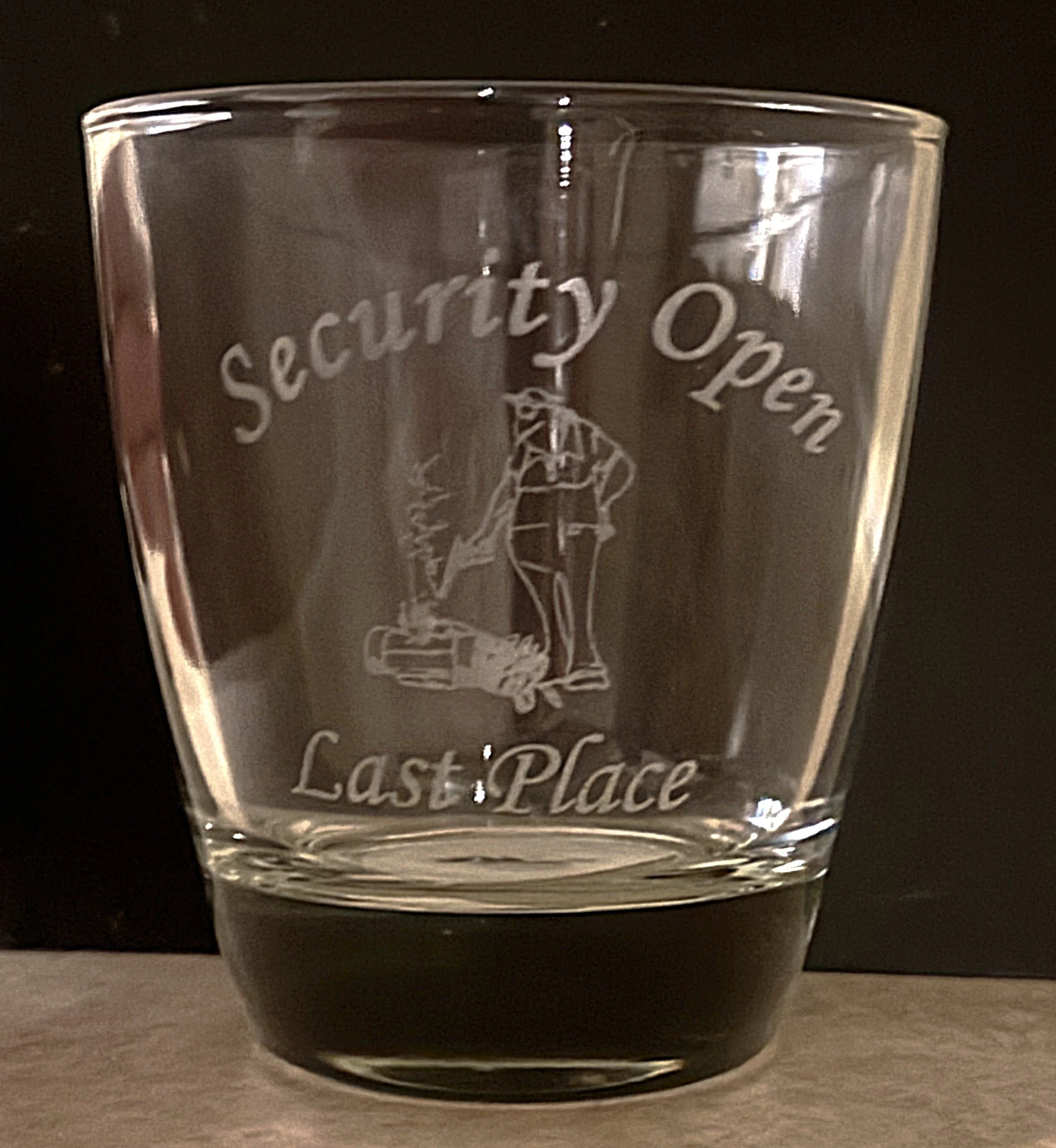 A clear glass with etched text and image that reads 'Security Open Last Place.' The etching shows a person at a desk or counter, with one arm resting on the surface.