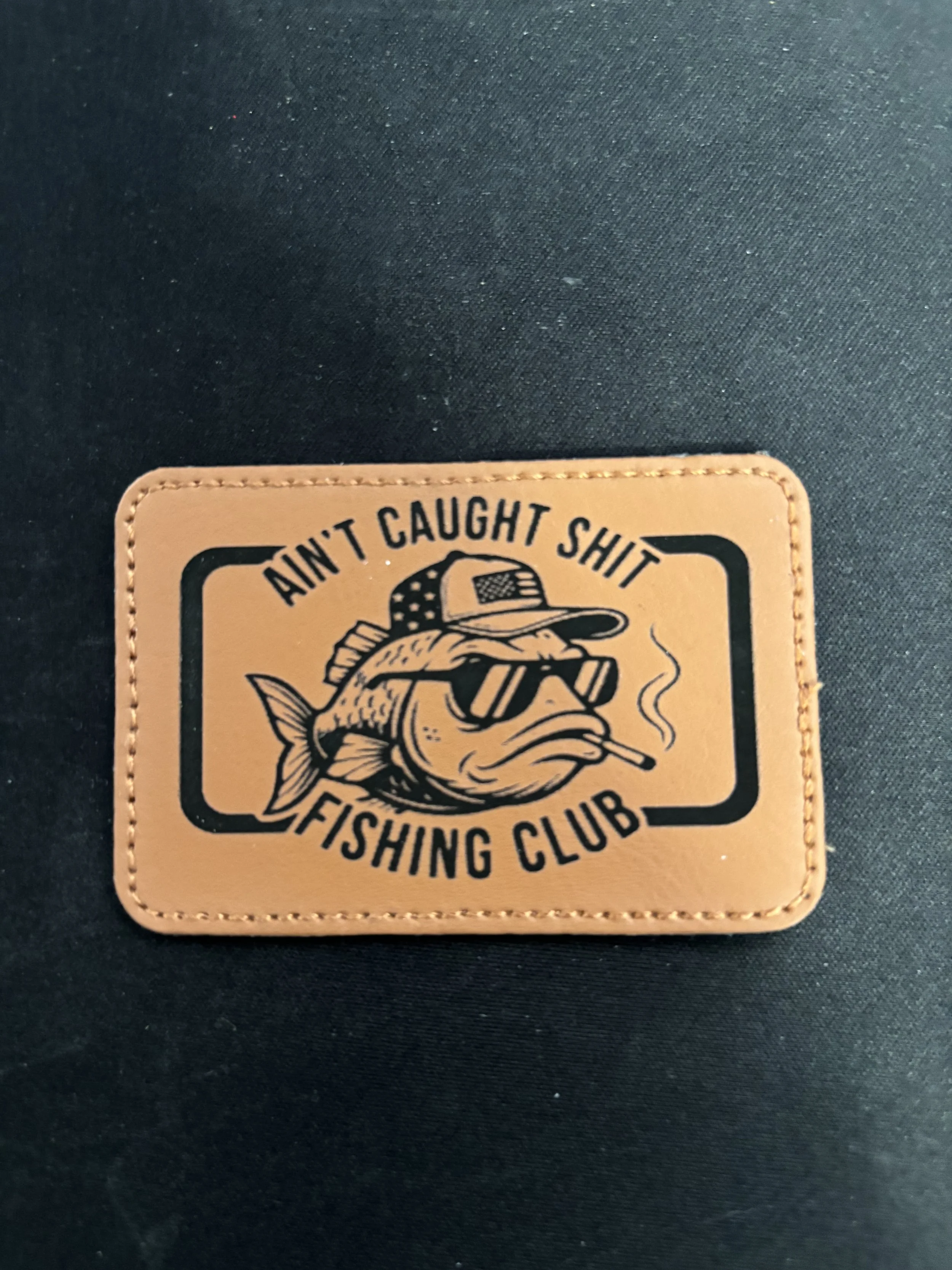 A patch with a fish wearing sunglasses, a cap, and a cigarette in its mouth, with the text 'Ain't Caught Shit Fishing Club' around it.