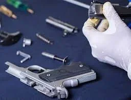 A person wearing a white glove is handling a small part near a disassembled semi-automatic pistol on a blue surface, with various gun components and tools around.