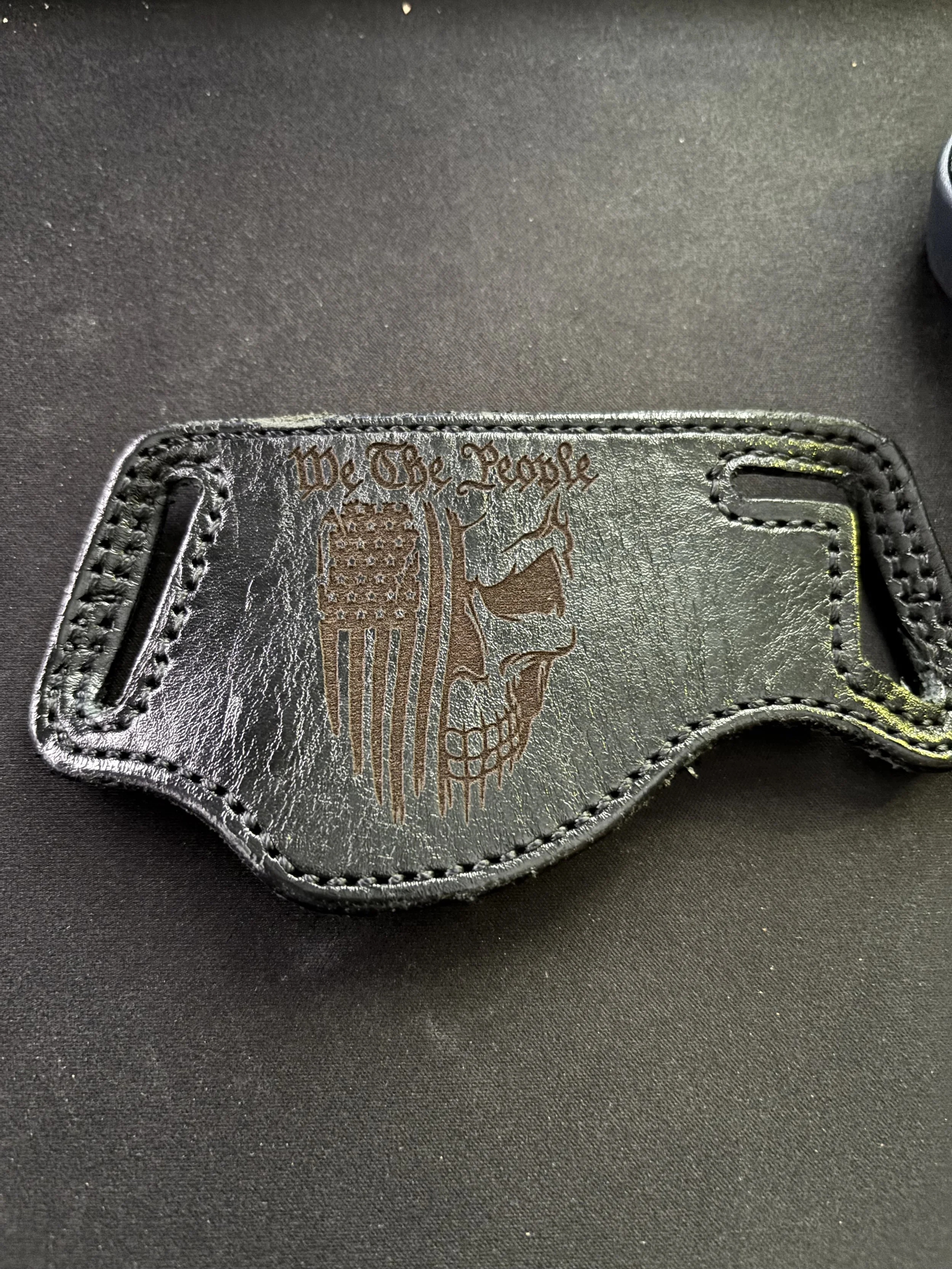 Black leather object with an engraved American flag and skull design, along with the text 'We The People' engraved above.