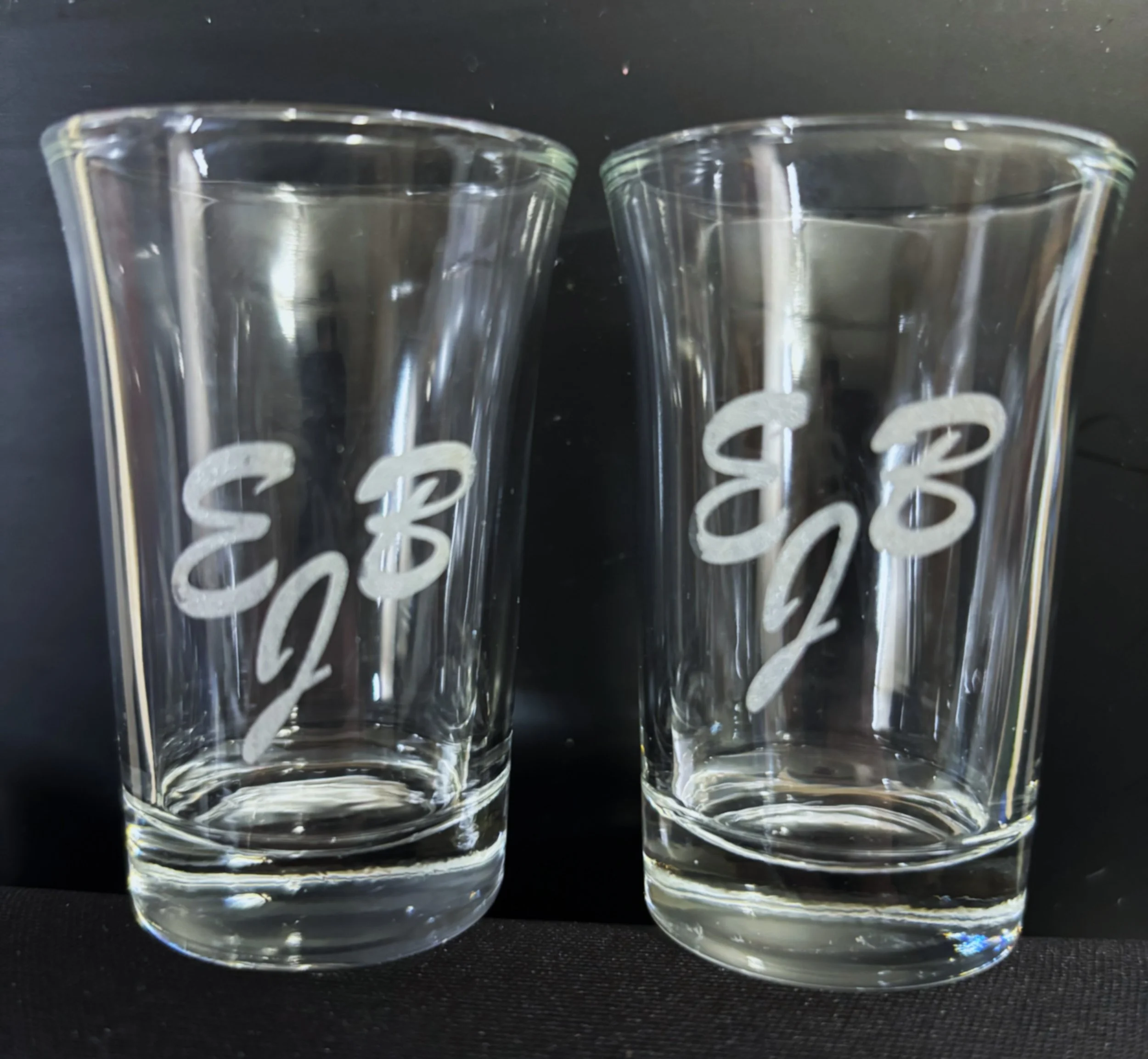 Two clear glass tumblers with white etched lettering that reads '8 2/3'. The tumblers are placed side by side on a black surface with a black background.