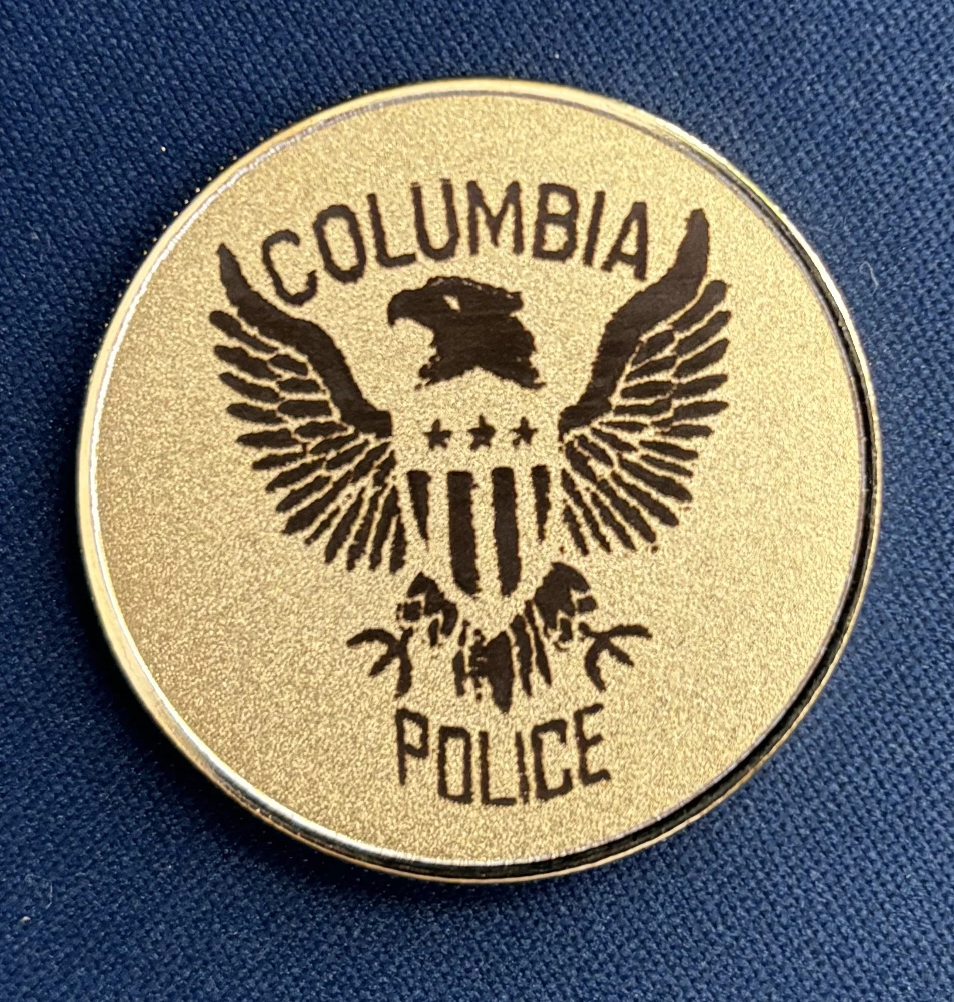 Gold-colored Columbia Police badge with a black eagle emblem and the words 'Columbia Police' on a dark blue fabric background.