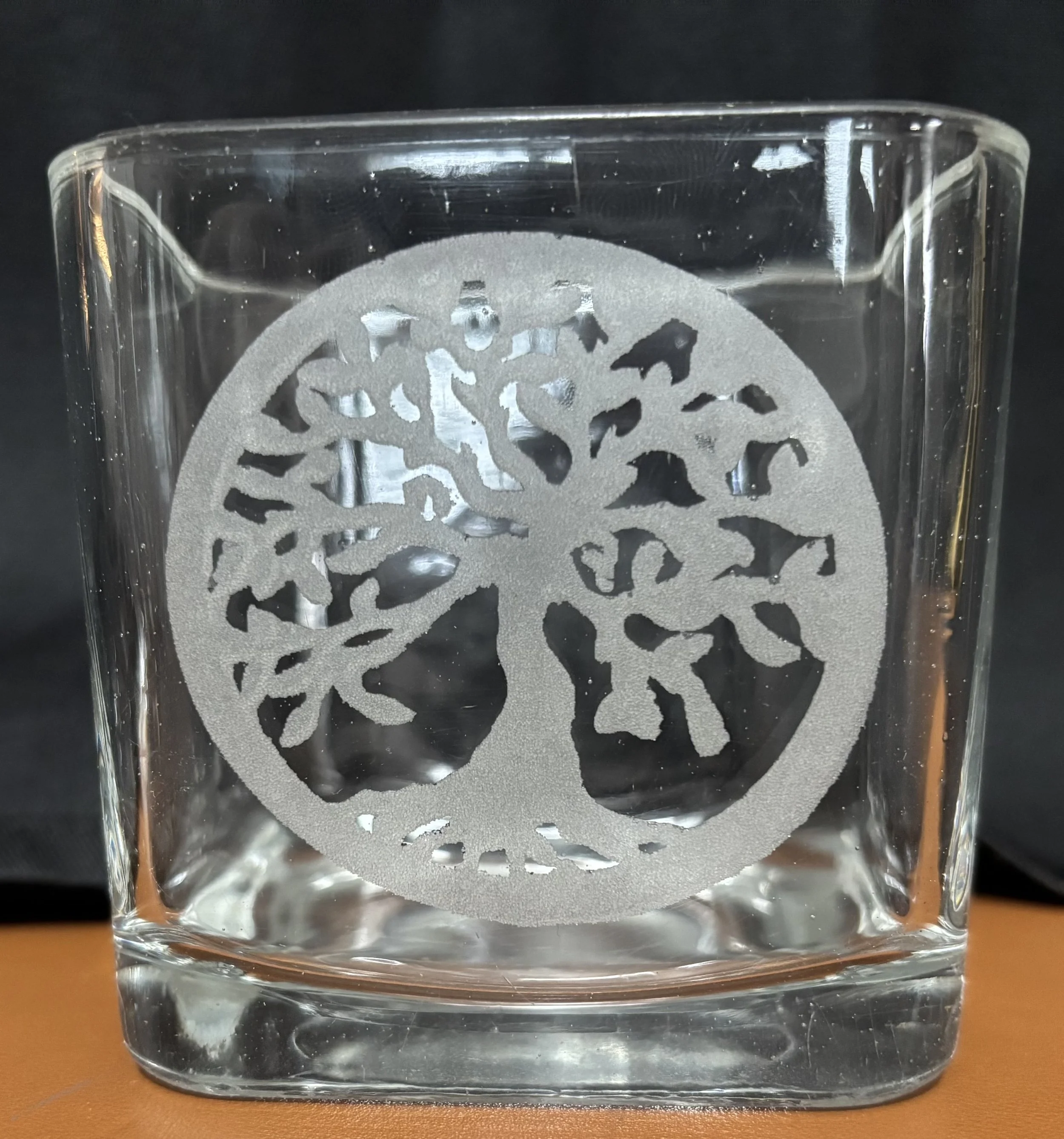 A clear glass with an etched design of a tree inside a circle, set against a black background.