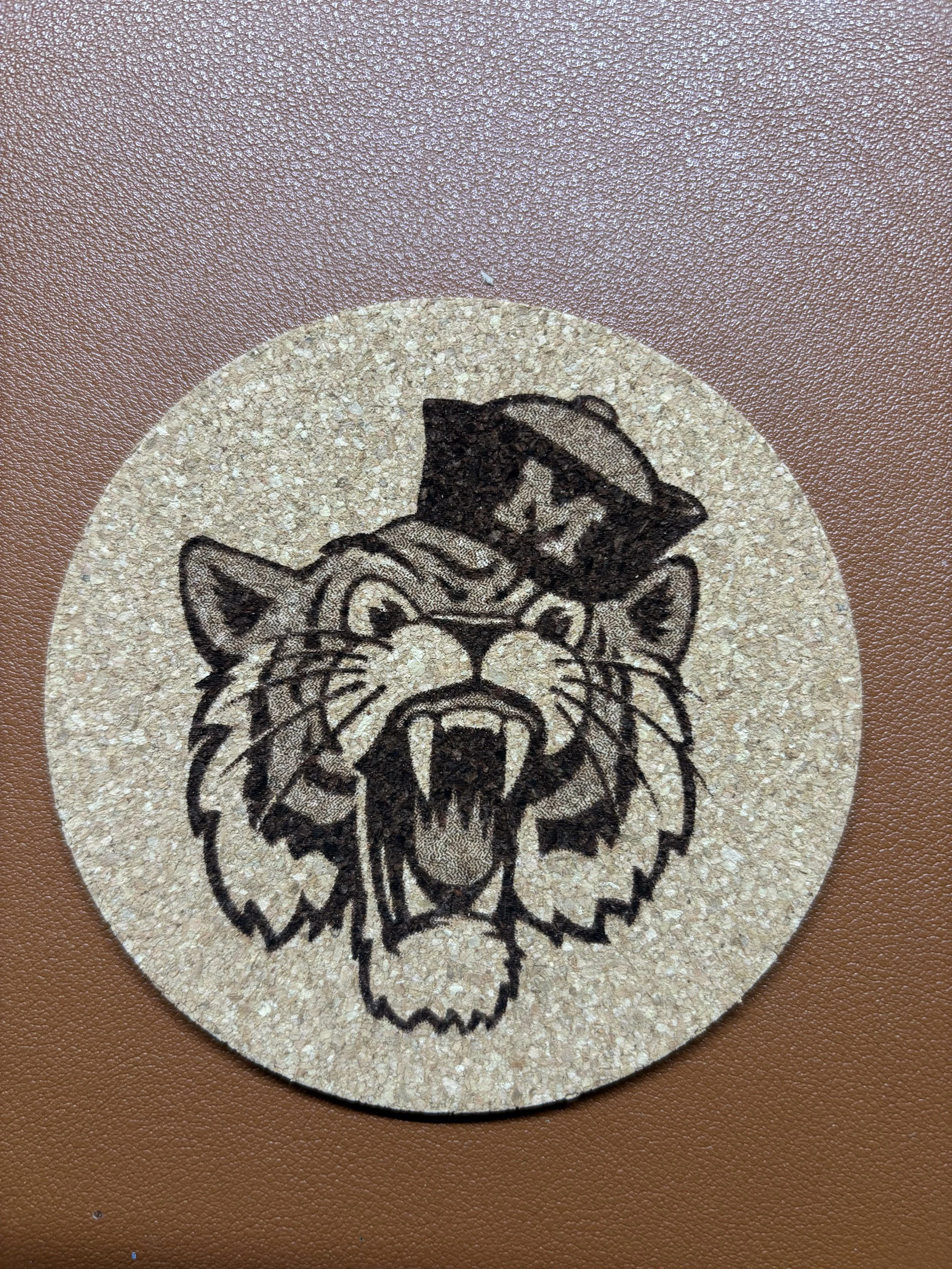 A round sticker featuring a wildcat wearing a graduation cap, outlined in black on a beige background, placed on a brown surface.