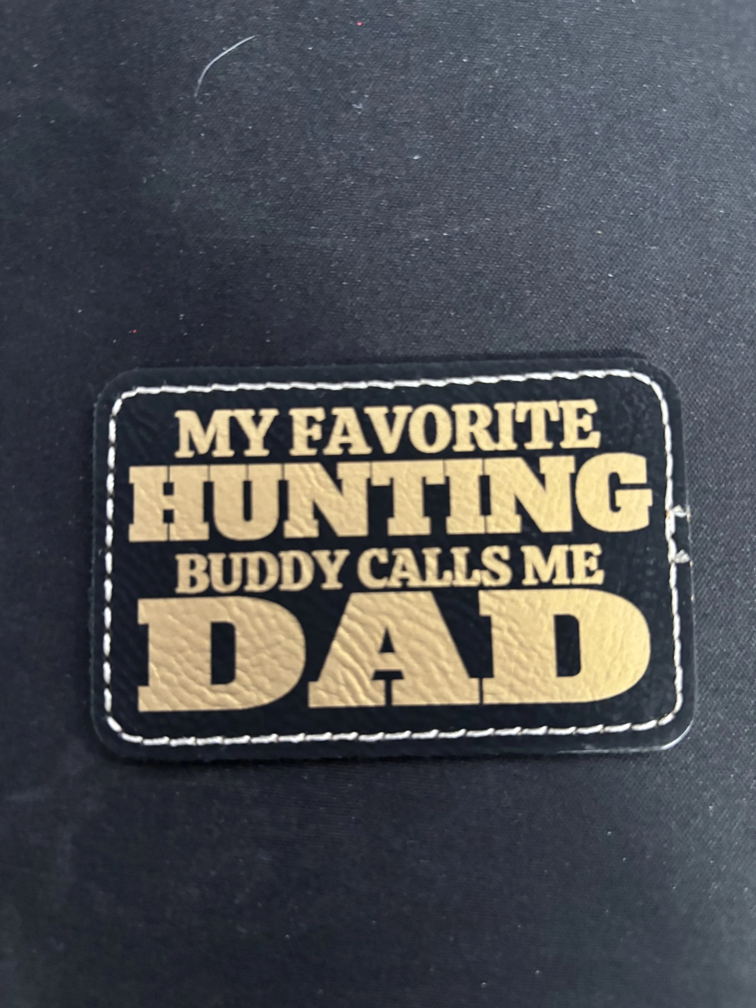 A patch with the text 'My Favorite Hunting Buddy Calls Me Dad' stitched on a black fabric background.