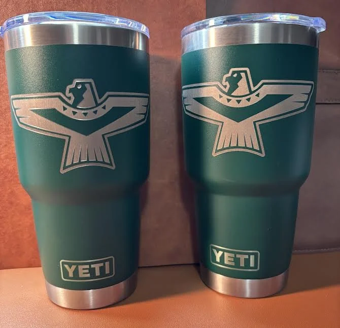 Two green YETI travel tumblers with a silver eagle logo and a silver base, placed on a wooden surface.