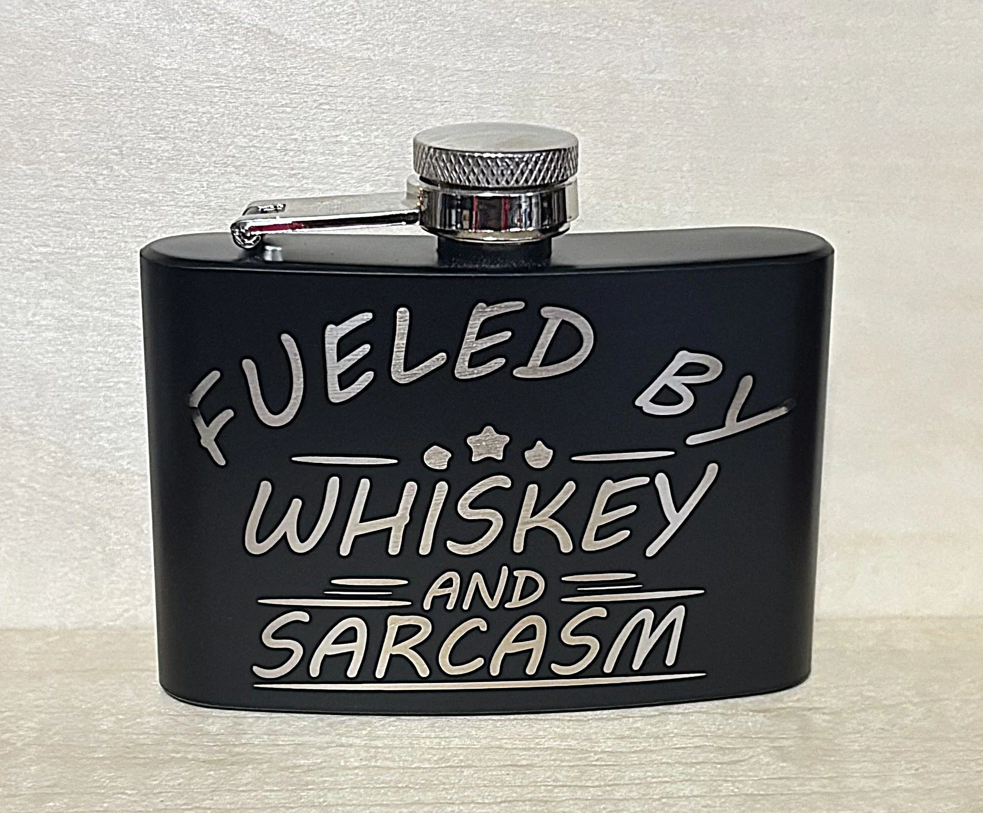 Black hip flask with silver text reading "Fueled by Whiskey and Sarcasm"