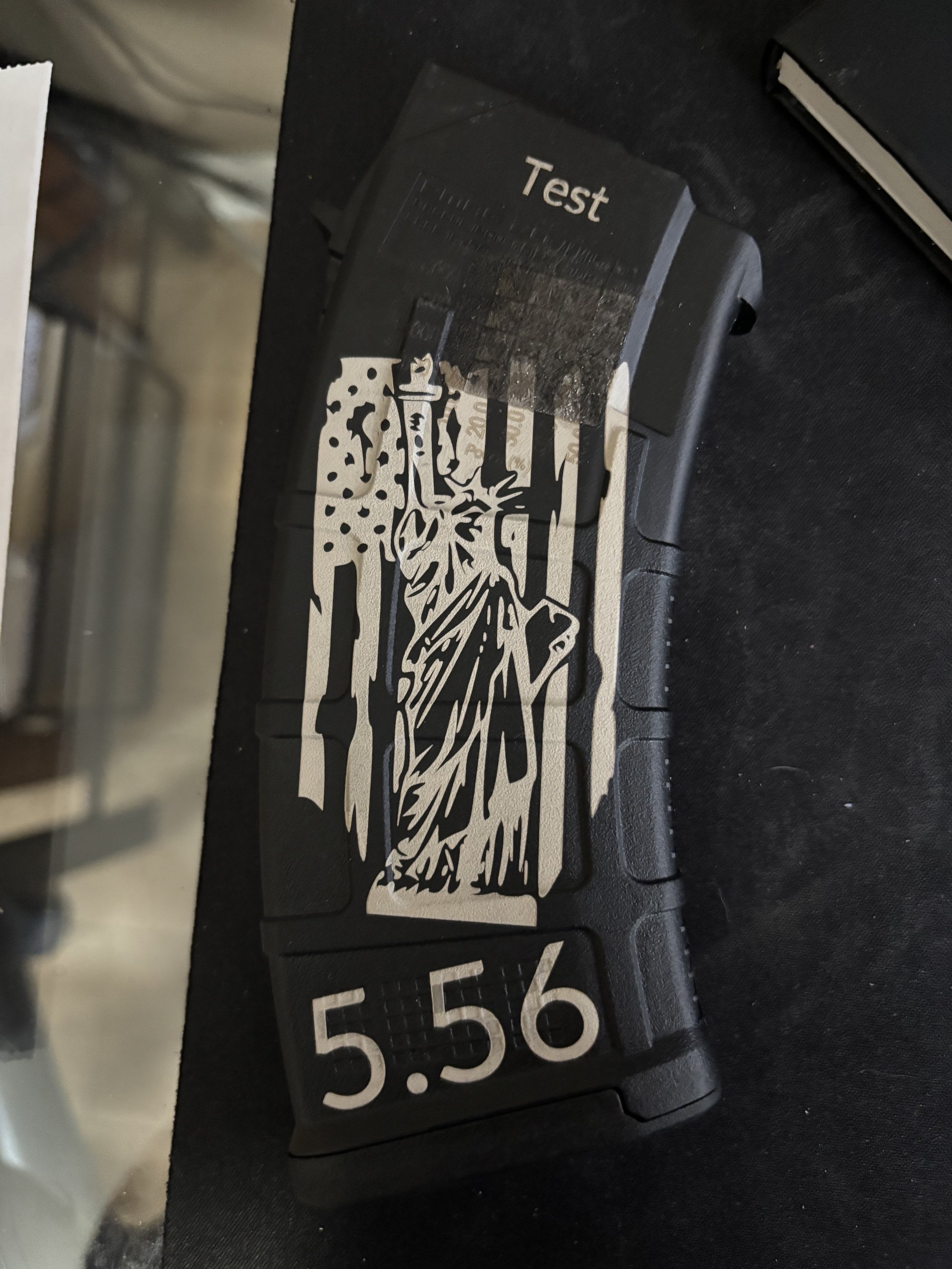 A black device with a sticker of the Statue of Liberty and a bullet proof vest depiction, displaying the numbers '5.56' and a testing label.
