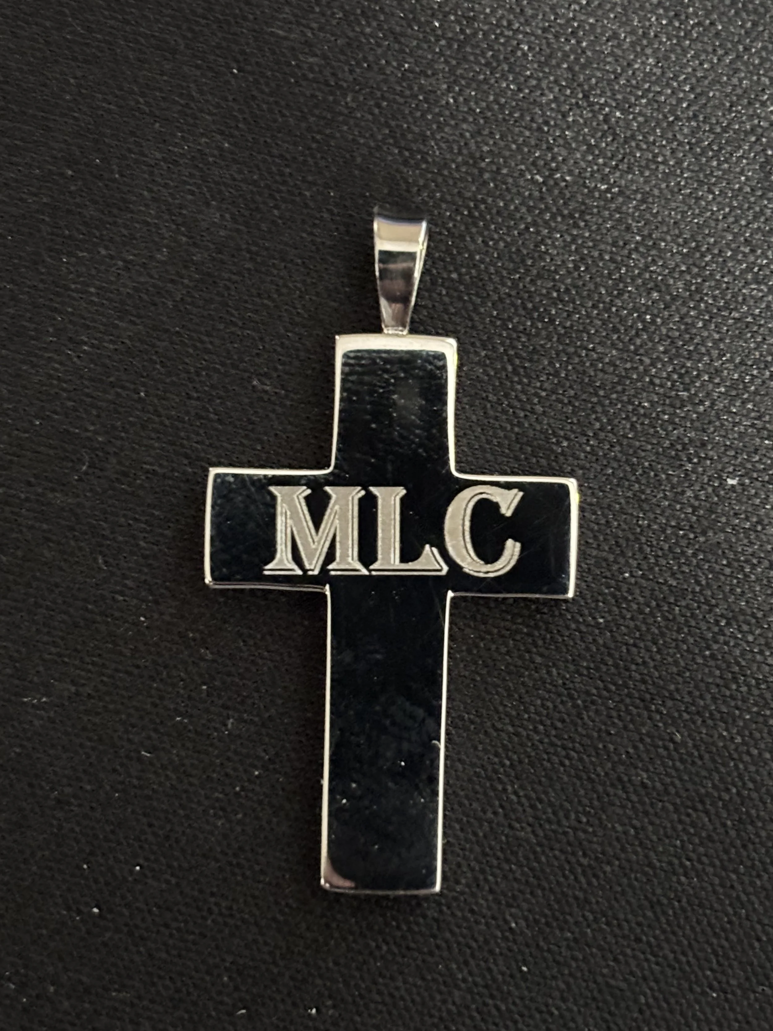 A silver cross pendant with the letters "MLC" engraved in the center, placed on a black textured surface.