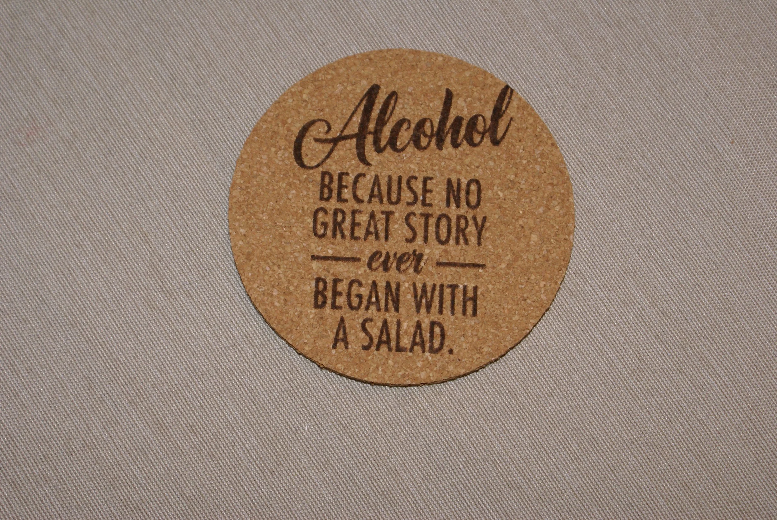 A round cork coaster with the quote, "Alcohol because no great story ever began with a salad," written on it, placed on a gray textured surface.