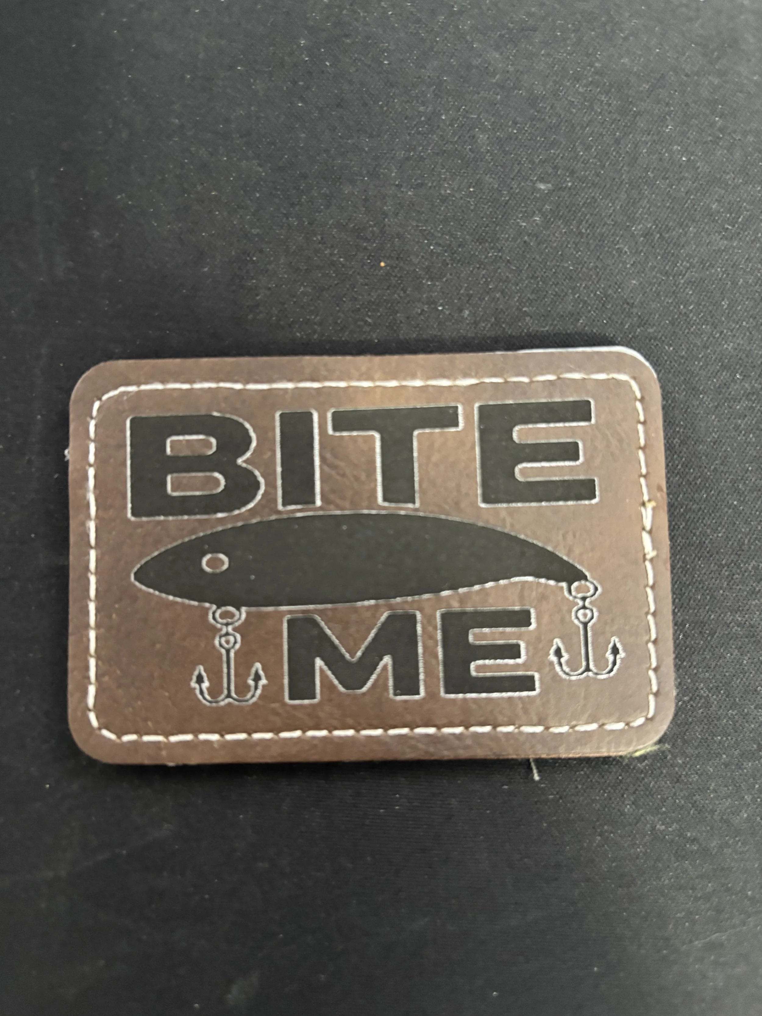 A brown leather patch with black and white embroidered text that reads 'BITE ME,' with a fishing lure and hook design.