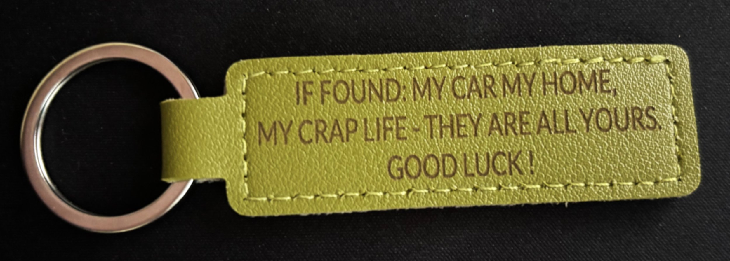 A yellow leather keychain with a metal ring and inscription that reads, "IF FOUND: MY CAR MY HOME MY CRAP LIFE & THEY ARE ALL YOURS GOOD LUCK!" on a black background.