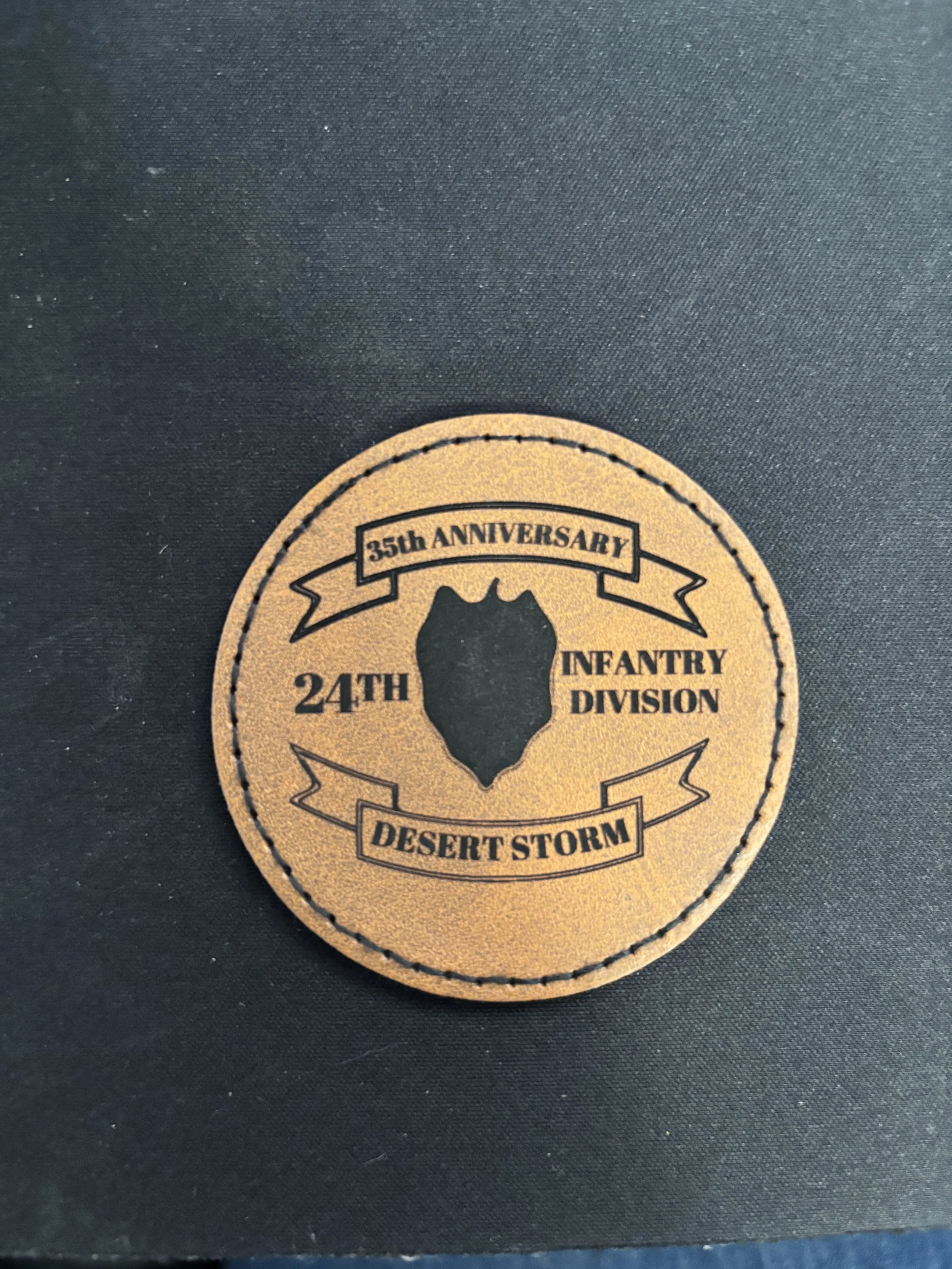 A circular patch with a tan background and black text and graphics. It features a central silhouette of a shield, with text around it that reads "35th Anniversary," "24th Infantry Division," and "Desert Storm."