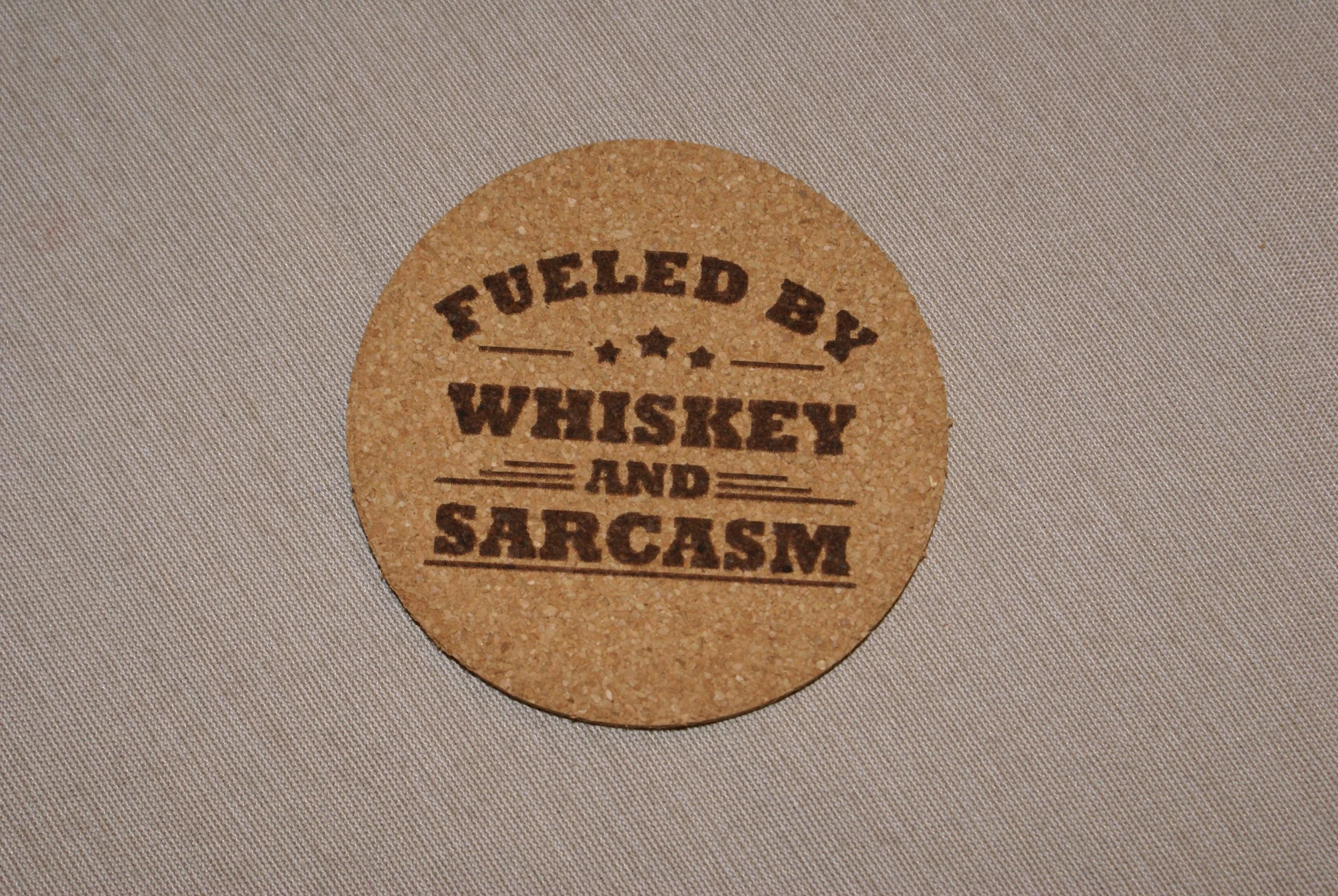 A circular cork coaster with the text "Fueled by Whiskey and Sarcasm" printed in dark brown.