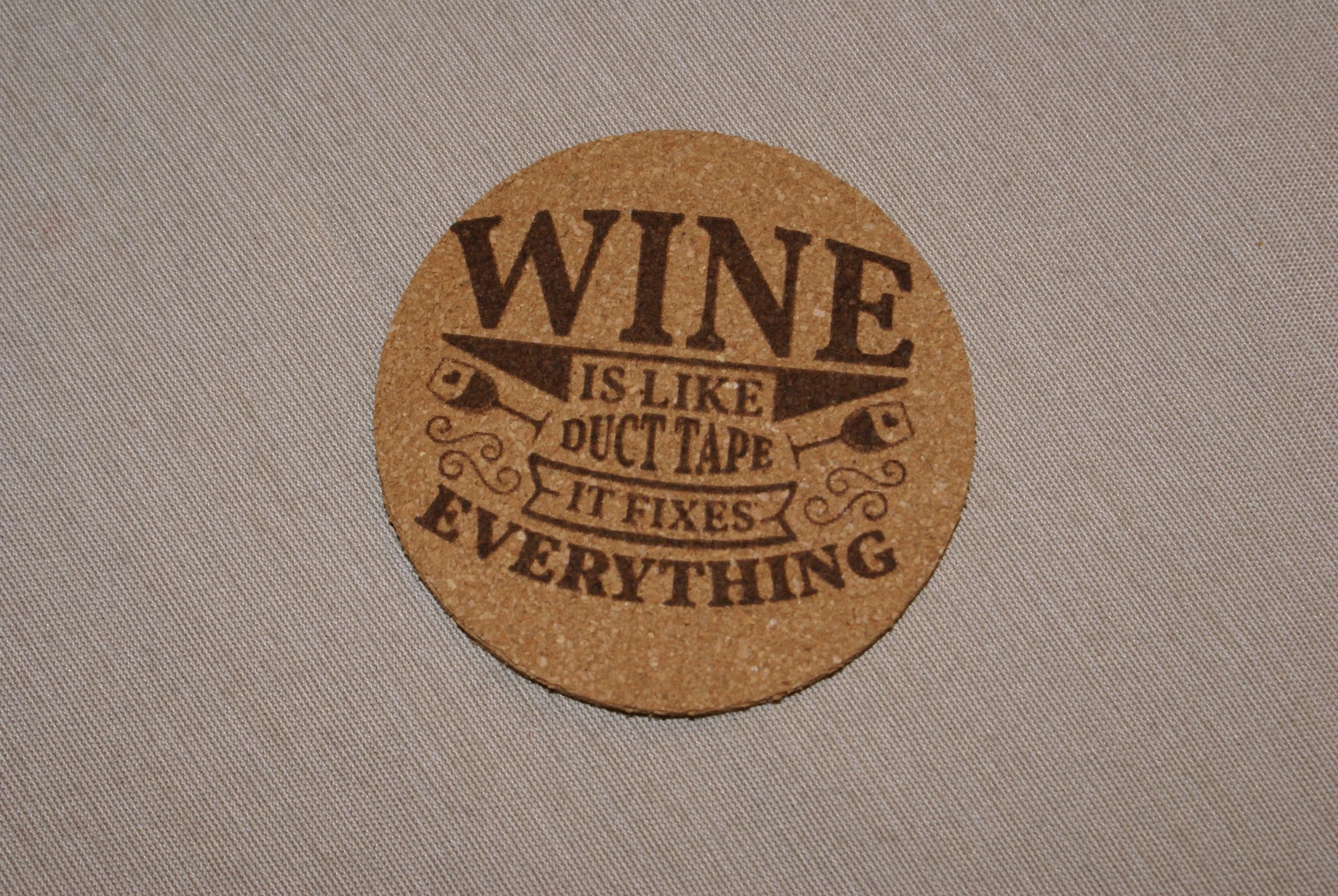 A round cork coaster with the text 'WINE IS LIKE DUCT TAPE IT FIXES EVERYTHING' printed in black.