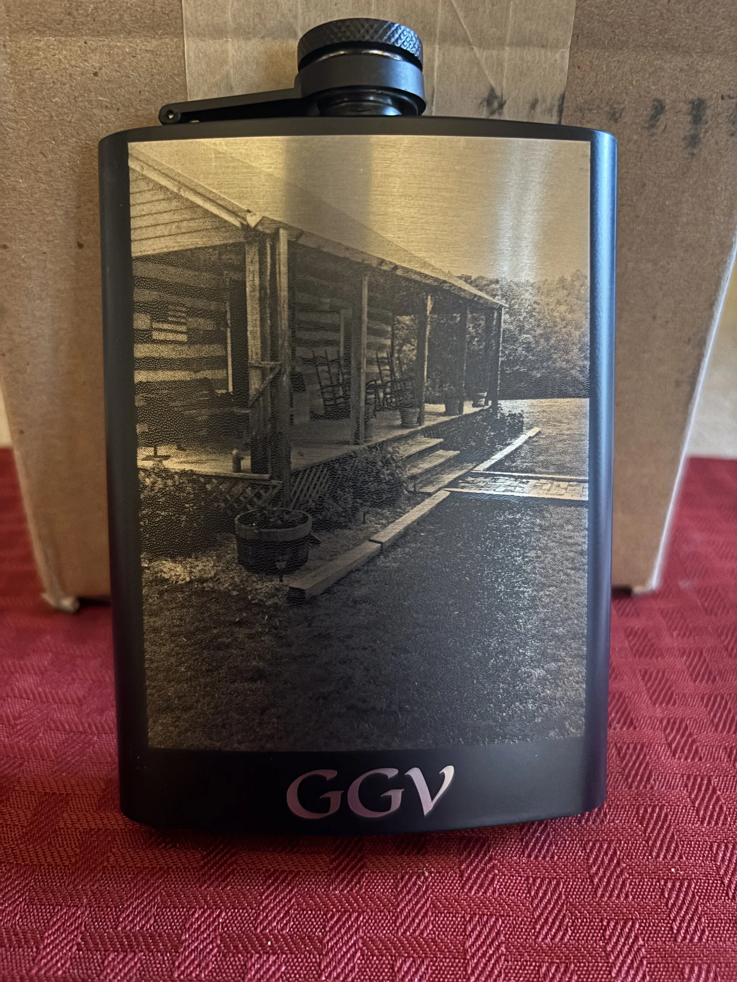 Black flask with a sepia-toned photo of a porch with steps and chairs printed on it, placed on a red textured surface.