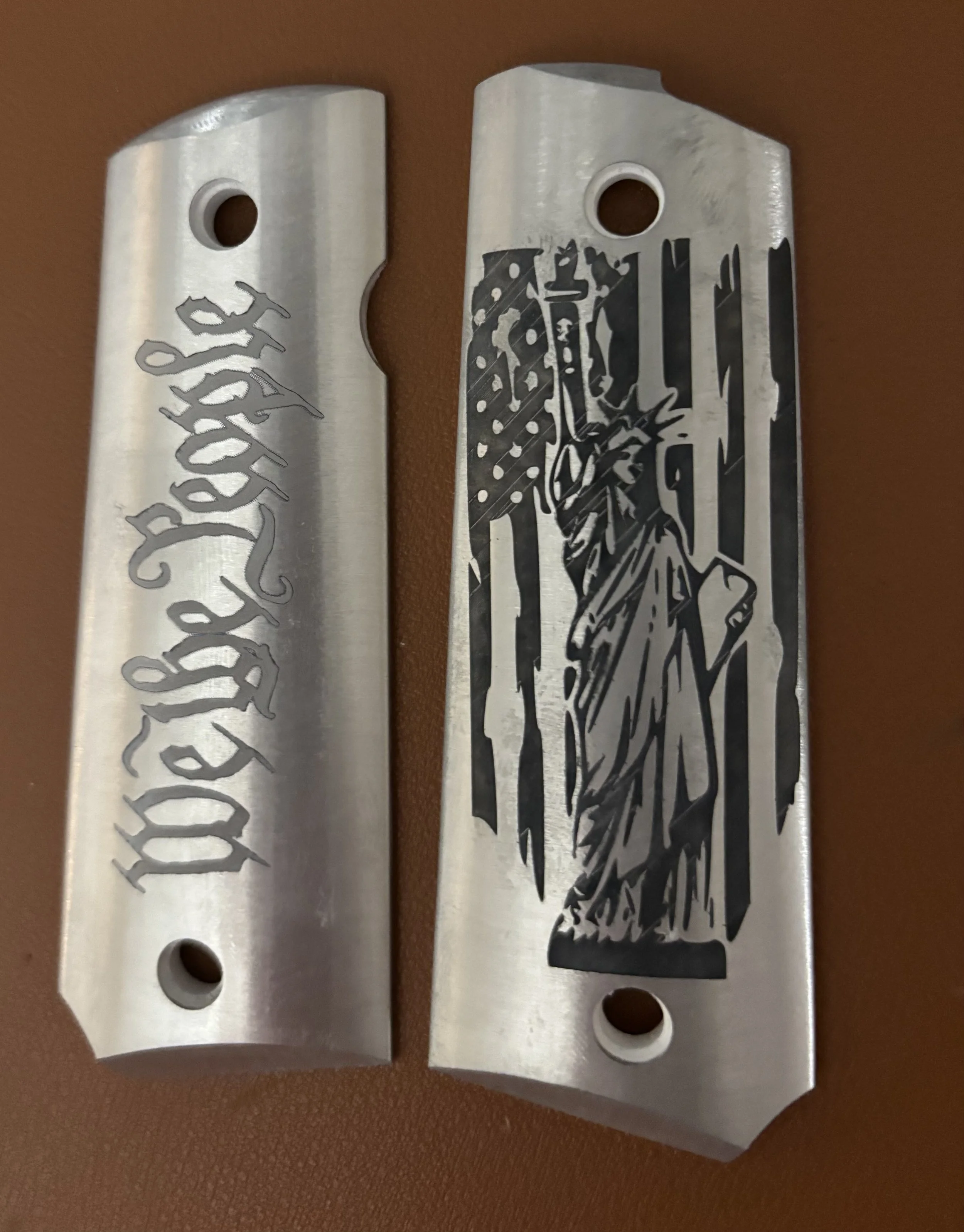 Two metallic gun grips with engraved designs, one with gothic text, the other with an illustration of the Statue of Liberty.