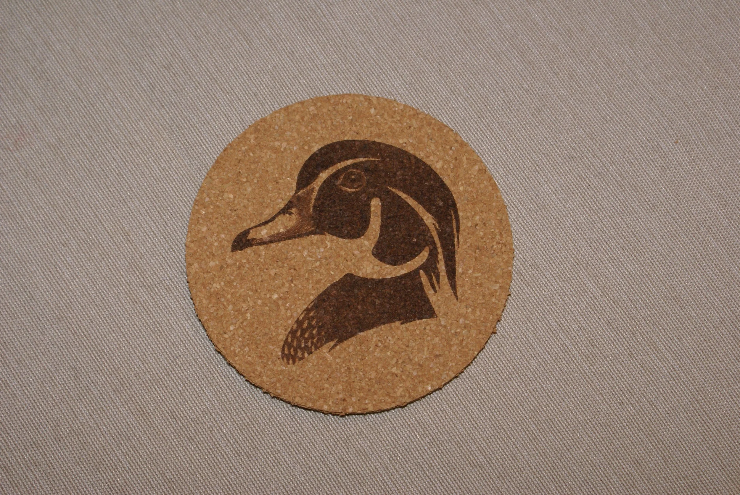 Close-up of a round cork coaster with a black silhouette of a duck's head on a white surface.