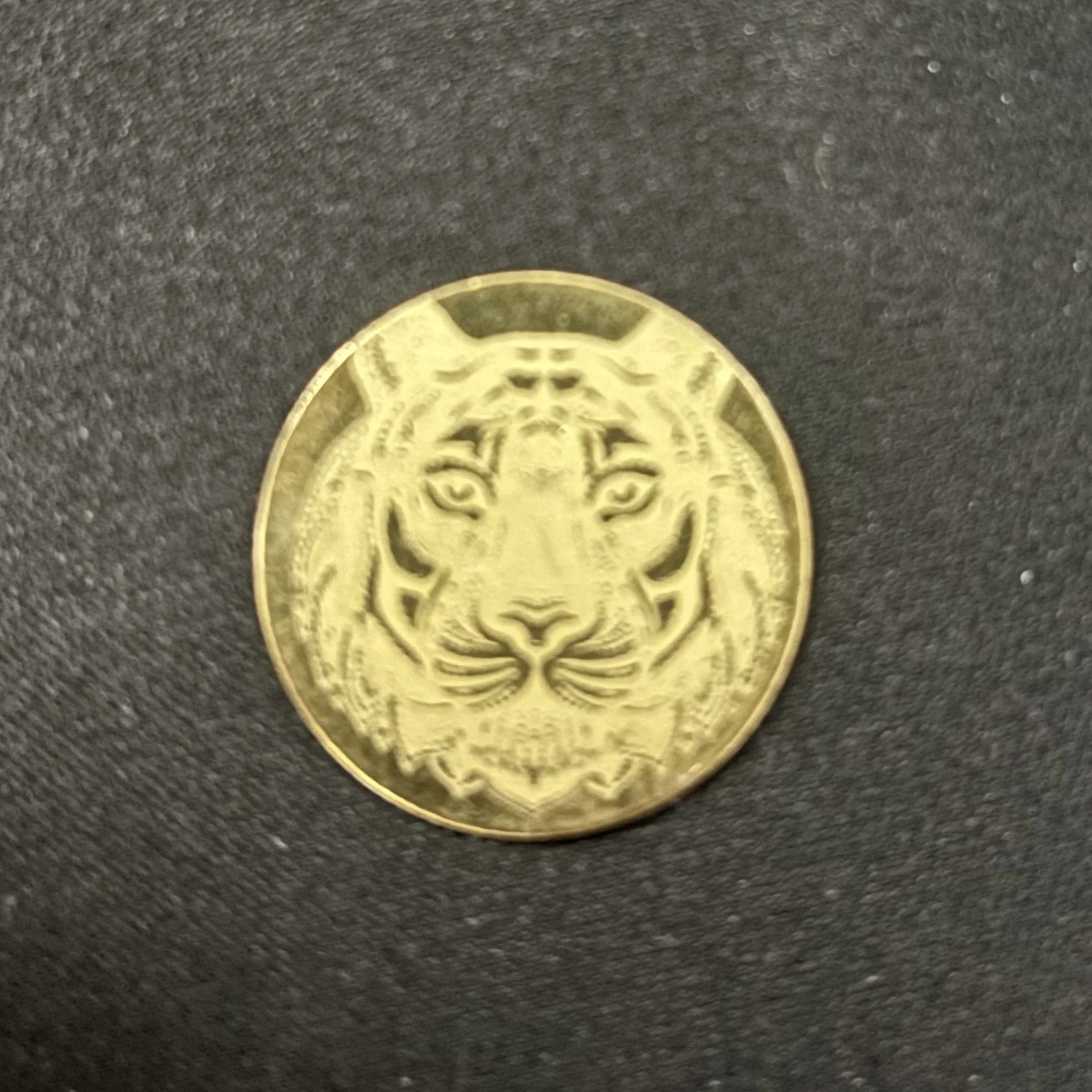 A gold coin with a tiger's face engraved on it, placed on a dark textured surface.