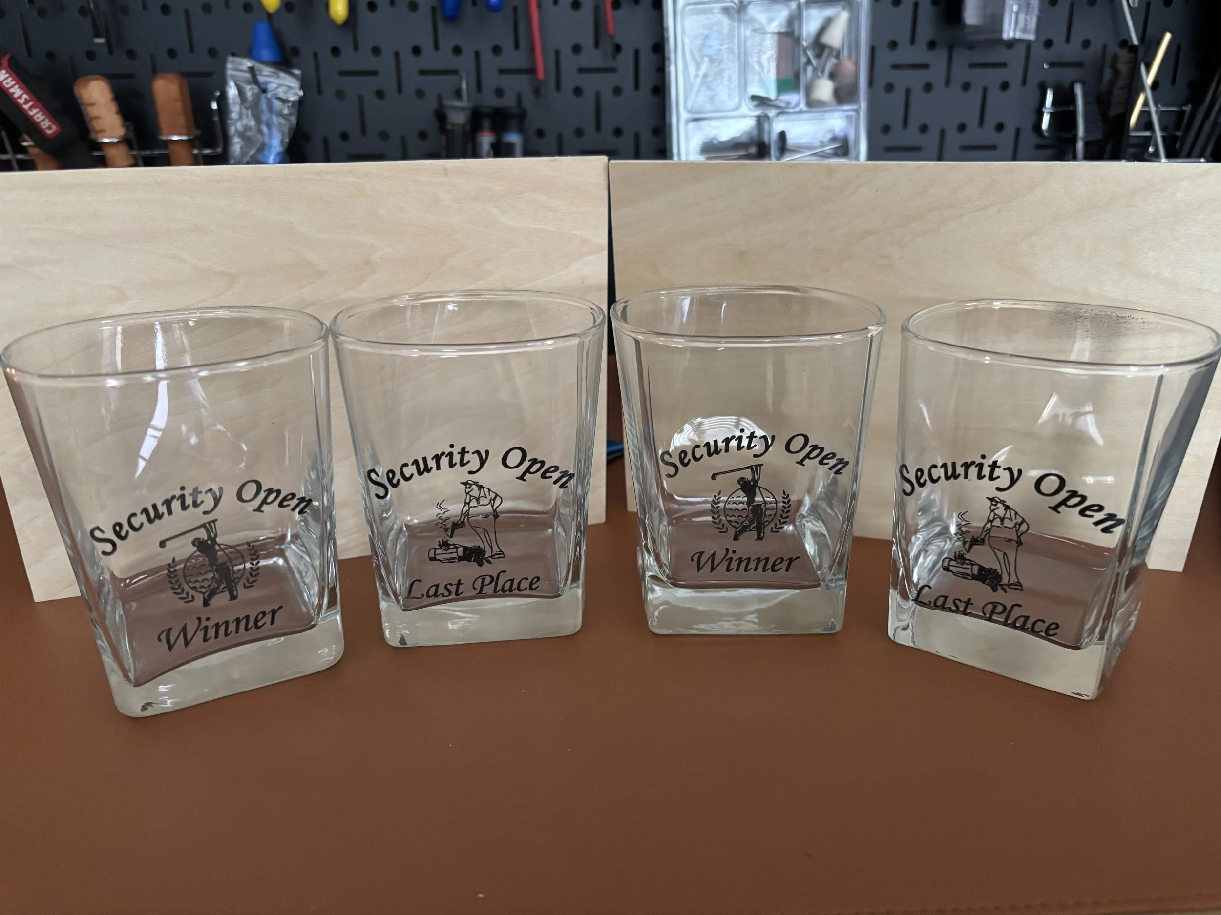 Four clear glass tumblers with black text and a graphic of a man with a beer mug, arranged on a brown surface in front of a wooden backdrop with a black pegboard and tools hanging.