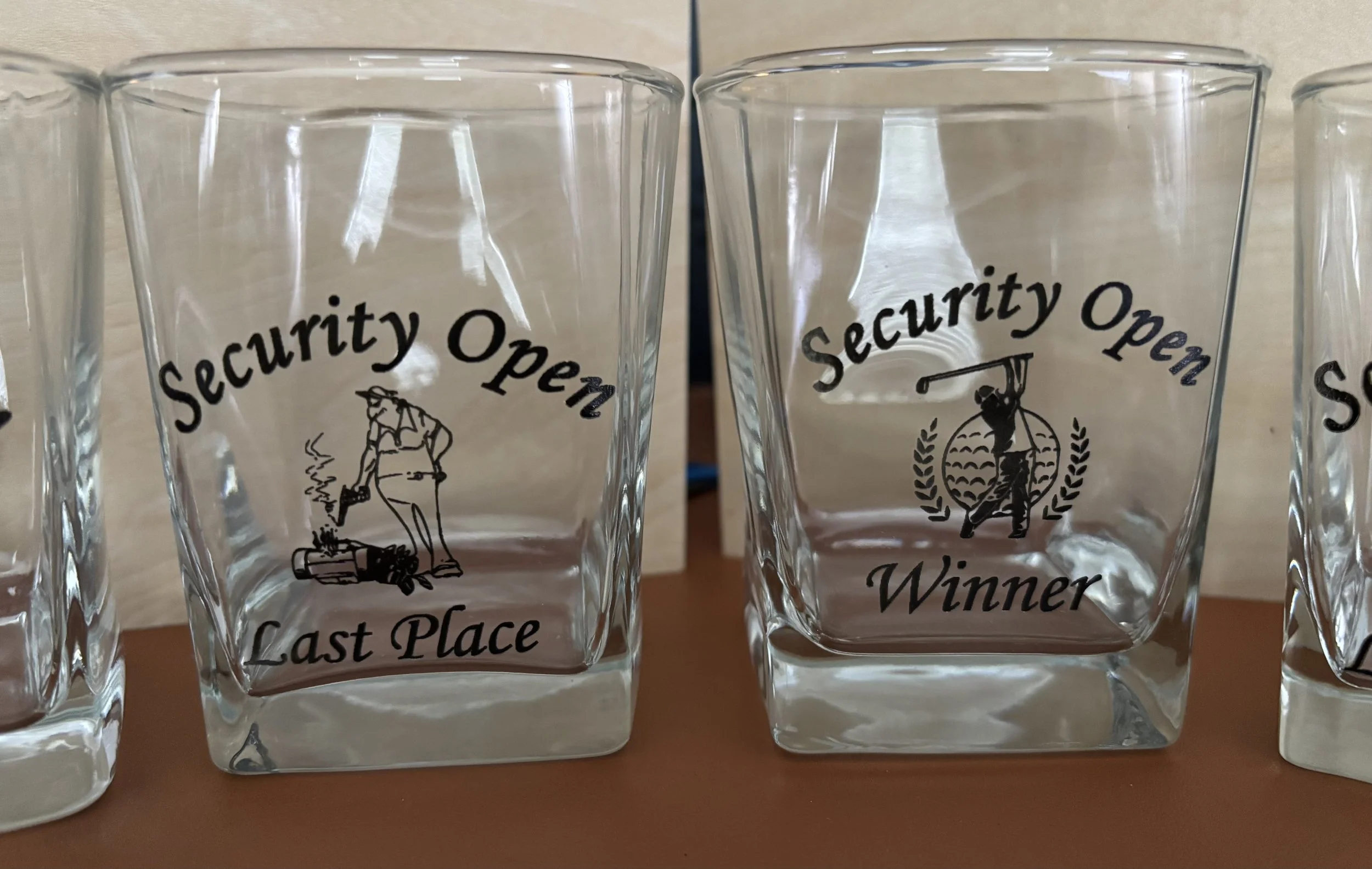 Two glass trophies with black text and illustrations. The left trophy says 'Security Open Last Place' and features a person in a trench coat and hat probing the ground with a magnifying glass. The right trophy says 'Security Open Winner' and depicts 