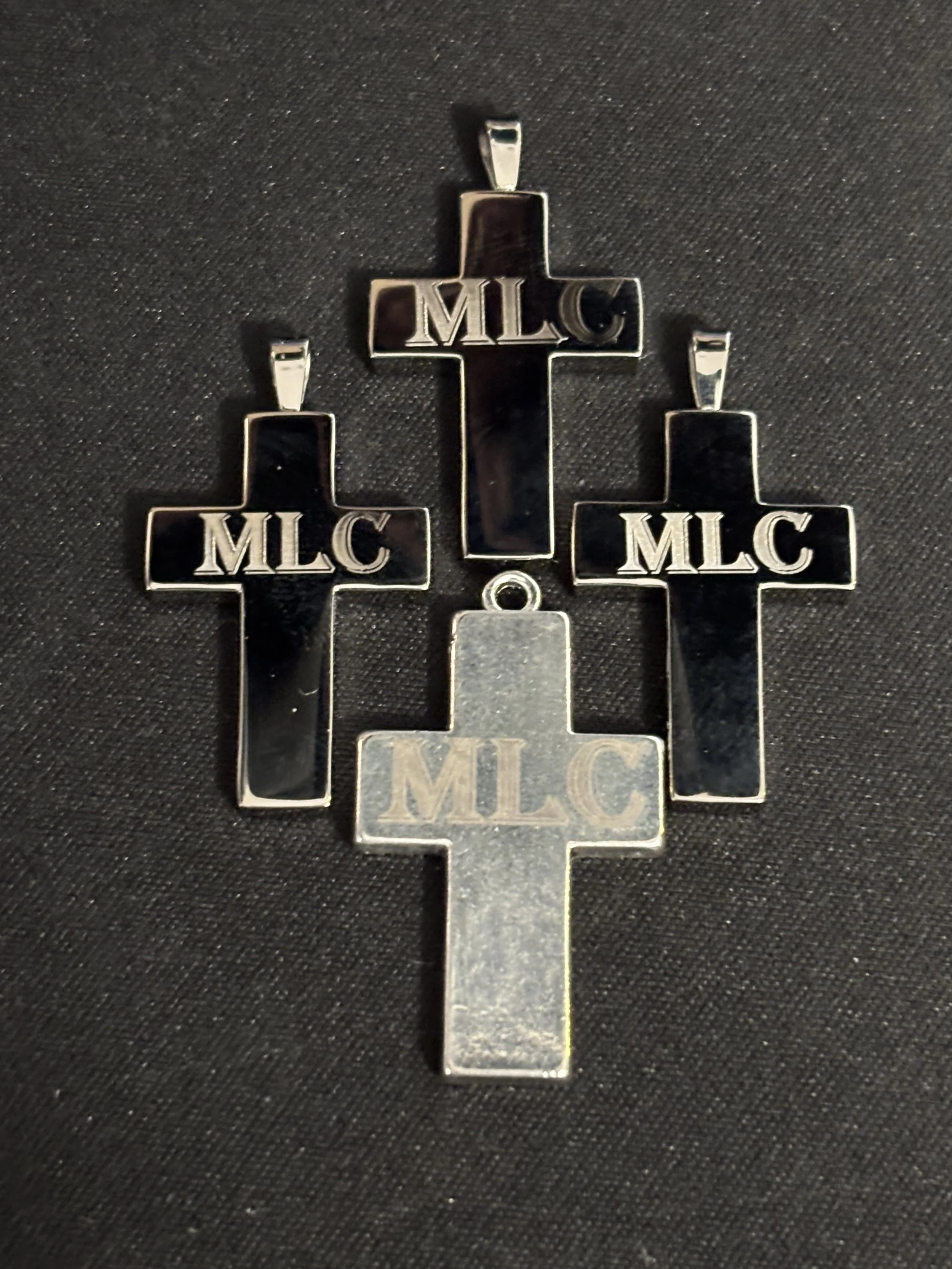 Four silver Christian crosses with the letters 'MLC' engraved on each, laid on a black surface.