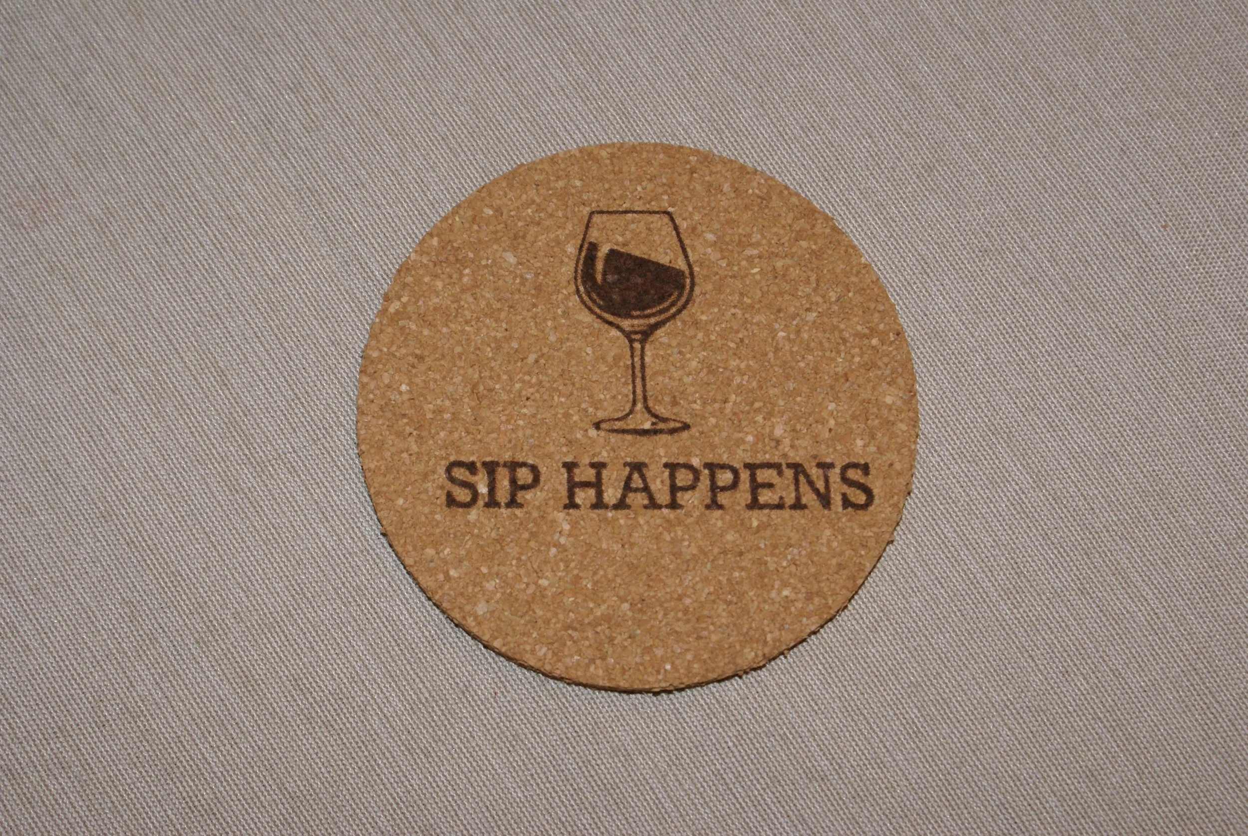 A round cork coaster with a wine glass illustration and the phrase 'SIP HAPPENS' written underneath.