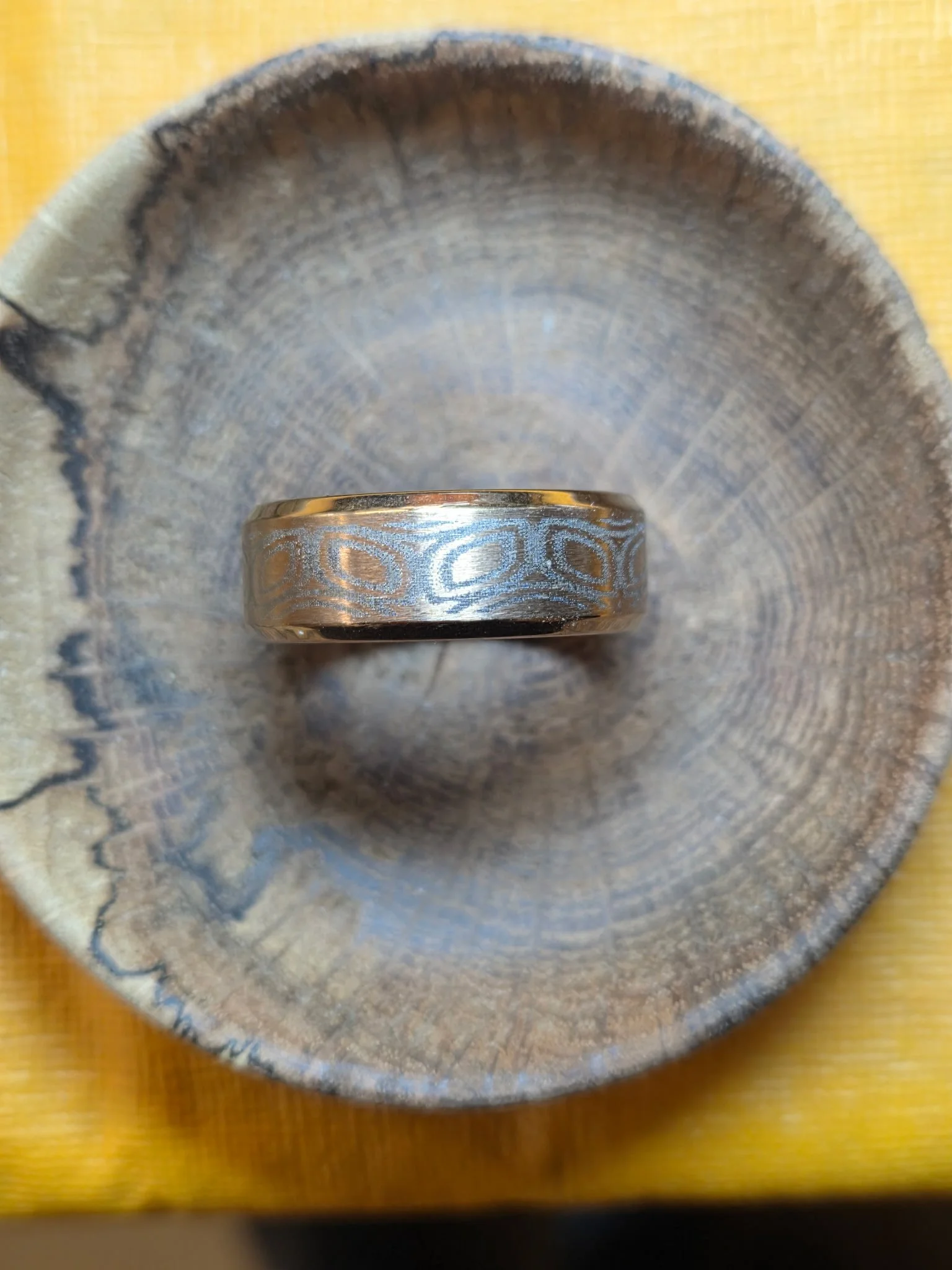 A silver wedding band with engraved circular patterns, placed inside a hollowed-out piece of wood on a yellow surface.