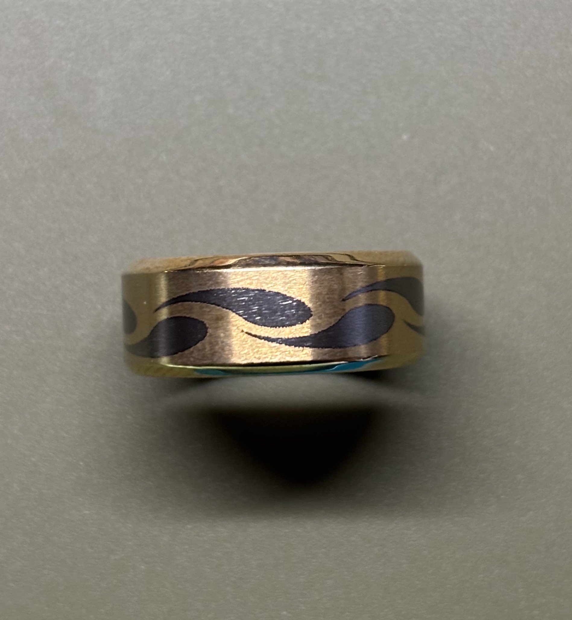 A close-up of a metallic ring with black flame-like designs on a light grey background.
